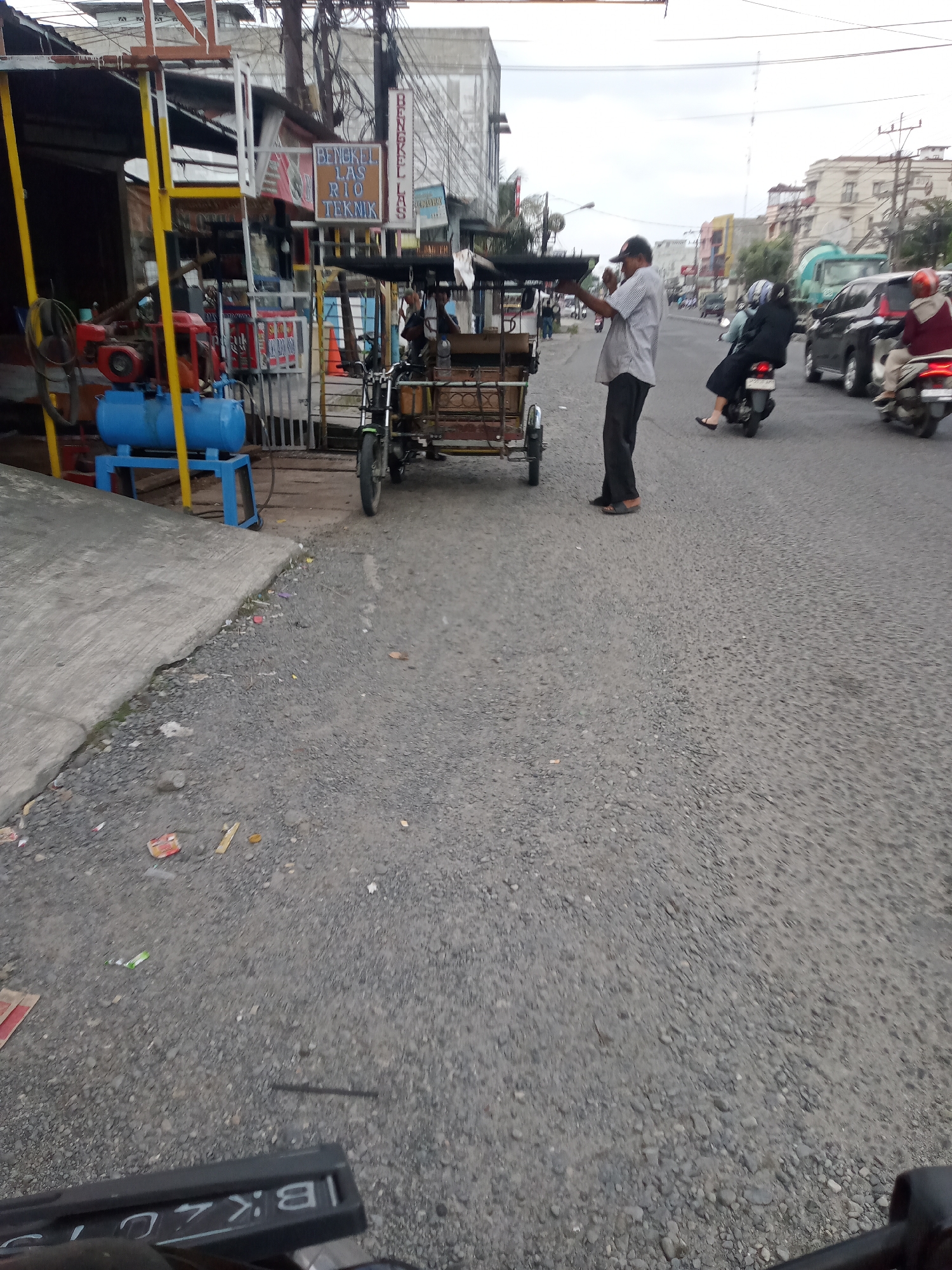 In the city of Sumber Melati Diski, Indonesia, a street scene unfolds with elements of commerce and traffic. A man in a light-colored striped shirt and dark pants stands near a three-wheeled vehicle, possibly a pedicab or cart, which is parked on the side of the road. He appears to be holding a cup or container. Behind him, a woman in dark clothing and a headscarf rides on the back of a motorcycle, and another person on a motorcycle, wearing a red and white jacket, is visible further down the street. Several other vehicles, including cars and trucks, are also present on the road, indicating moderate traffic. The roadside features a shop or workshop with a blue air compressor and other equipment. The ground is covered in gravel and asphalt, and debris is scattered along the edge of the road. Buildings line the street in the background, suggesting a commercial or mixed-use area. The sky is overcast.