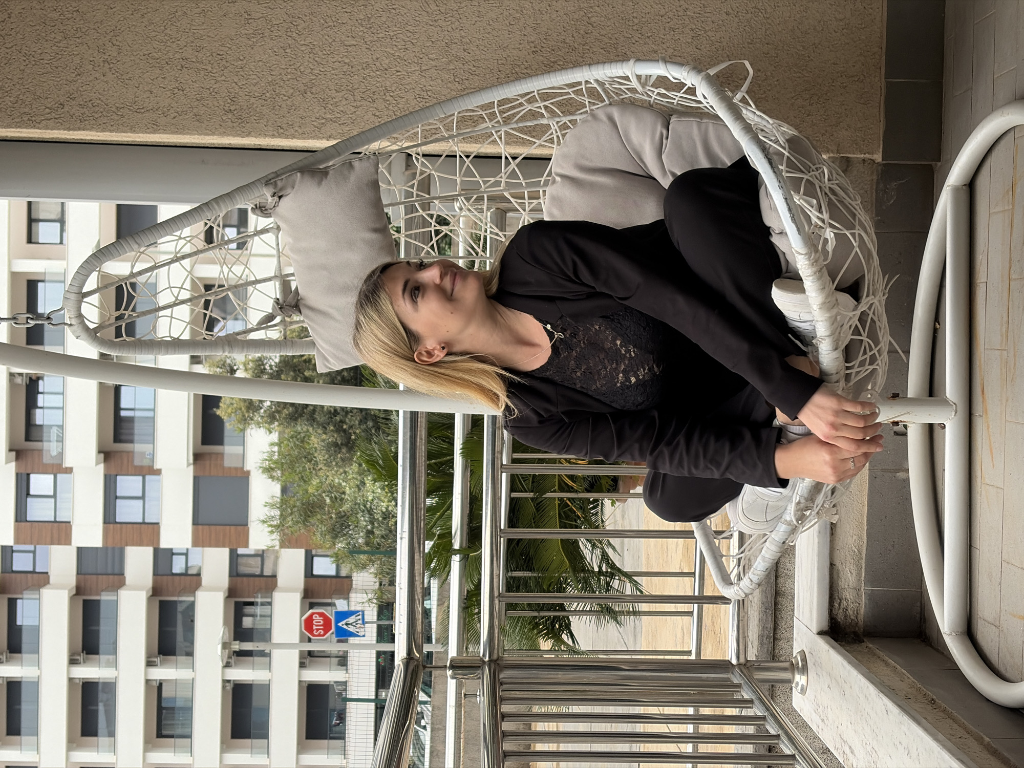 A woman with blonde hair, dressed in a black top with lace details, a black blazer, black trousers, and white sneakers, is sitting in a white hanging egg chair. Her body is angled slightly, and she is looking upwards and to her right with a relaxed expression, her hands resting on the chair's frame. The chair is furnished with light grey cushions and has a distinctive woven design.
The setting appears to be a balcony or terrace, with a metal railing in the foreground. The floor of the balcony is light-colored, possibly tiled. In the background, a modern apartment building with multiple balconies and dark-framed windows is visible. Lush green trees and foliage are also present, suggesting an outdoor, possibly urban or suburban, environment.
To the left of the frame, near the building, two road signs are visible. A red octagonal "STOP" sign is prominently displayed. Below it, a blue square sign with a white triangle containing a black pedestrian figure indicates a pedestrian crossing. The lighting suggests it is daytime, with clear visibility. The overall atmosphere is calm and relaxed. The architectural style and road signs are consistent with the location context of Budva, Montenegro.