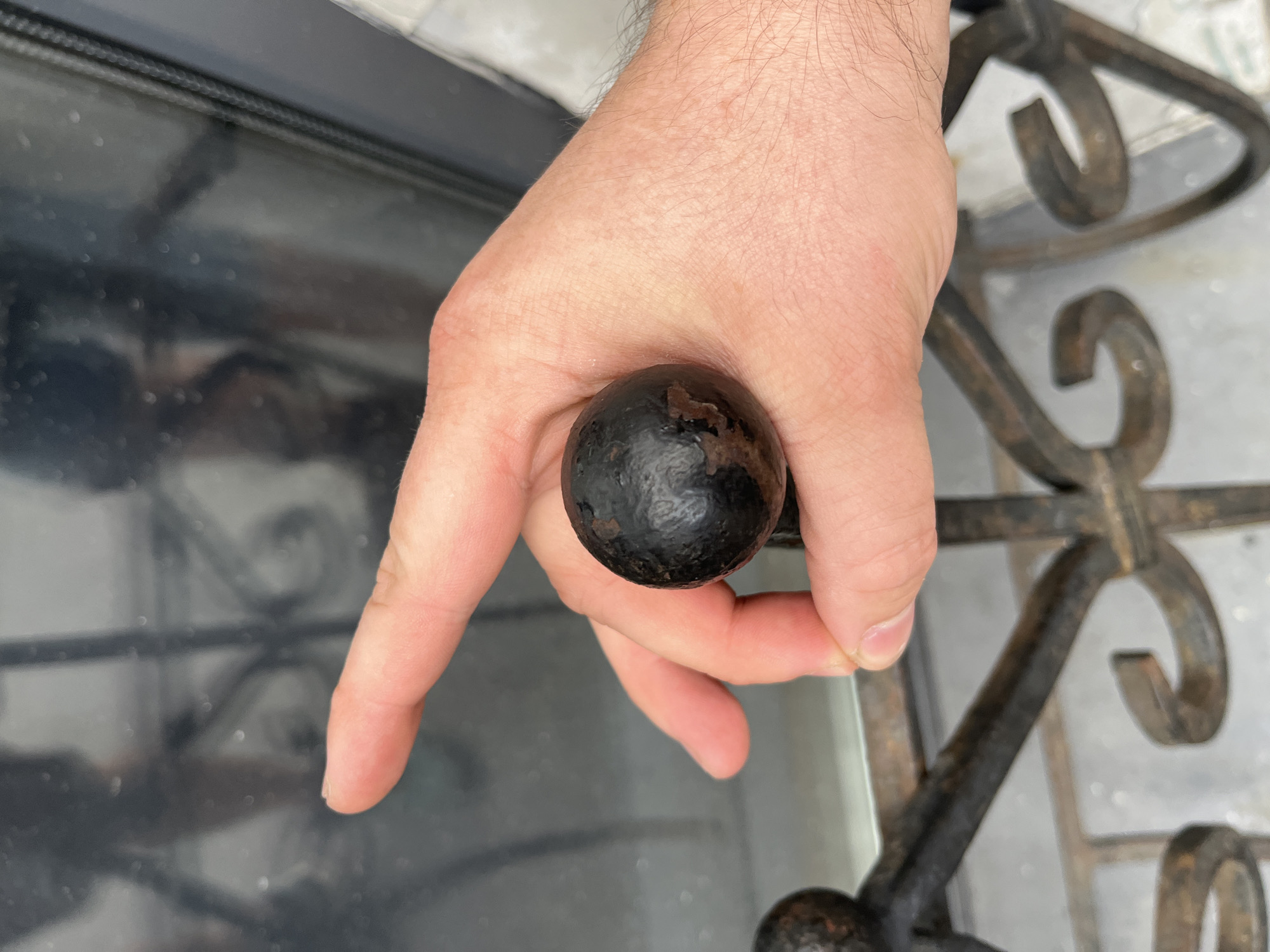 A hand holds a dark, spherical object, possibly an old cannonball, against a backdrop of a blurred cityscape. The object is textured with visible pitting and rust marks. The hand is positioned in the foreground, with fingers gently gripping the sphere. The background suggests an urban environment, with reflections of buildings and structures visible on a dark, possibly glass or polished surface. The presence of a wrought-iron railing in the lower part of the image hints at architectural details that could be found in historical districts. The overall impression is one of historical artifact discovered within a modern urban setting. The specific location in Saint Petersburg, Russia, cannot be definitively determined from the visual cues alone, but the architectural style of the railing and the general atmosphere could align with many of the city's historic areas.