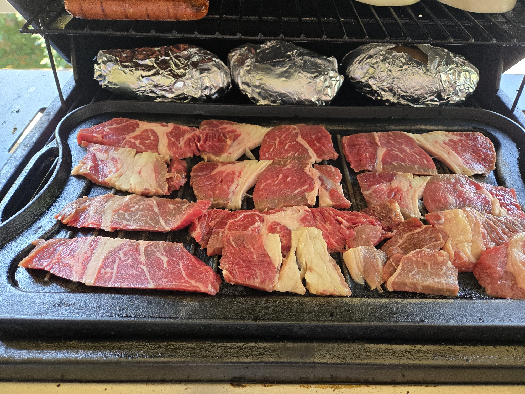 A close-up, high-angle, outdoor shot shows raw beef steaks arranged on a black griddle on a grill. The steaks vary in size and thickness, with visible marbling and fat. Behind the steaks, on the upper rack of the grill, are several items wrapped in aluminum foil, and a partially visible hot dog. The background is blurred, showing green foliage and hints of a patio or deck. The scene appears to be a domestic setting, likely a backyard barbecue. The specific location of Bellevue, United States of America, is not ascertainable from the visual information in the image.
