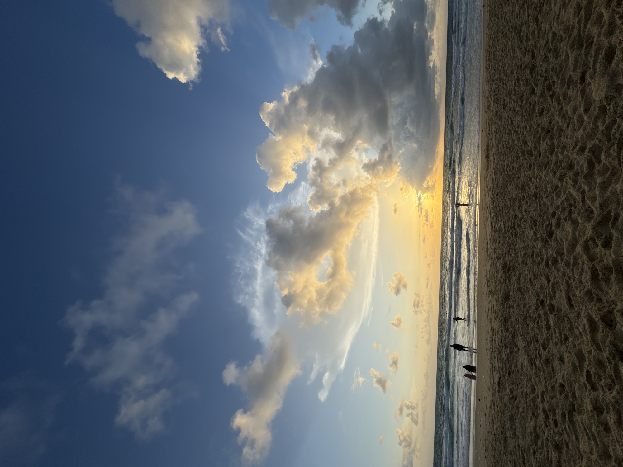 A wide-angle shot captures a tranquil beach scene at sunset. The foreground features coarse, rippled sand, leading to the edge of the ocean where gentle waves break. Several figures are visible in the shallow water and on the wet sand near the shoreline. On the far left, two individuals stand close together, with a pink object, possibly a surfboard or bodyboard, beside them. Further out, a solitary figure stands in the water. Towards the right, another lone figure is wading in the sea. The sky above is a dramatic canvas of blue, adorned with a variety of clouds. The sun, low on the horizon, casts a warm, golden glow through a break in the clouds, illuminating some of them with yellow and orange hues. The overall atmosphere is serene, with the natural beauty of the sky and sea dominating the scene. The image does not contain any identifying features of Karon, Thailand.