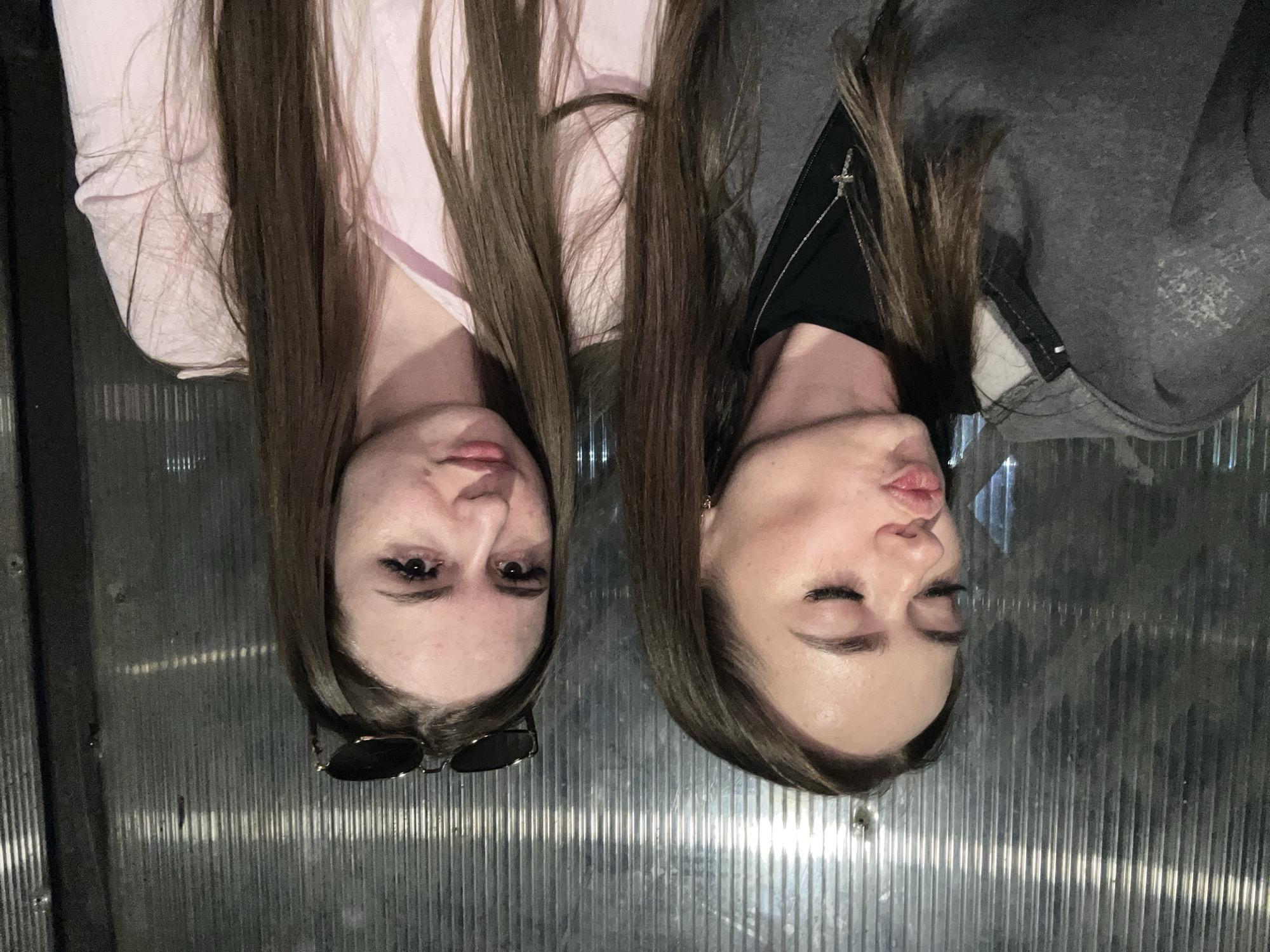 The image displays two young women, both with long, dark brown hair, positioned such that their heads are towards the bottom of the frame and their bodies extend upwards. The background consists of vertical, ribbed metallic panels that show some reflectivity, suggesting an indoor, possibly utilitarian or industrial setting.
The woman on the left wears a light pink top. Dark sunglasses are perched on top of her head. She gazes directly towards the viewer with a neutral expression.
The woman on the right is dressed in a dark top underneath a grey hooded jacket or sweatshirt. A silver chain with a small cross pendant is visible around her neck. She has her eyes closed and her lips pursed in a playful manner, as if blowing a kiss.
The overall activity appears to be a casual pose for a photograph, likely a selfie between friends. The lighting is indeterminate but suggests an indoor environment. There is no visible text in the image.