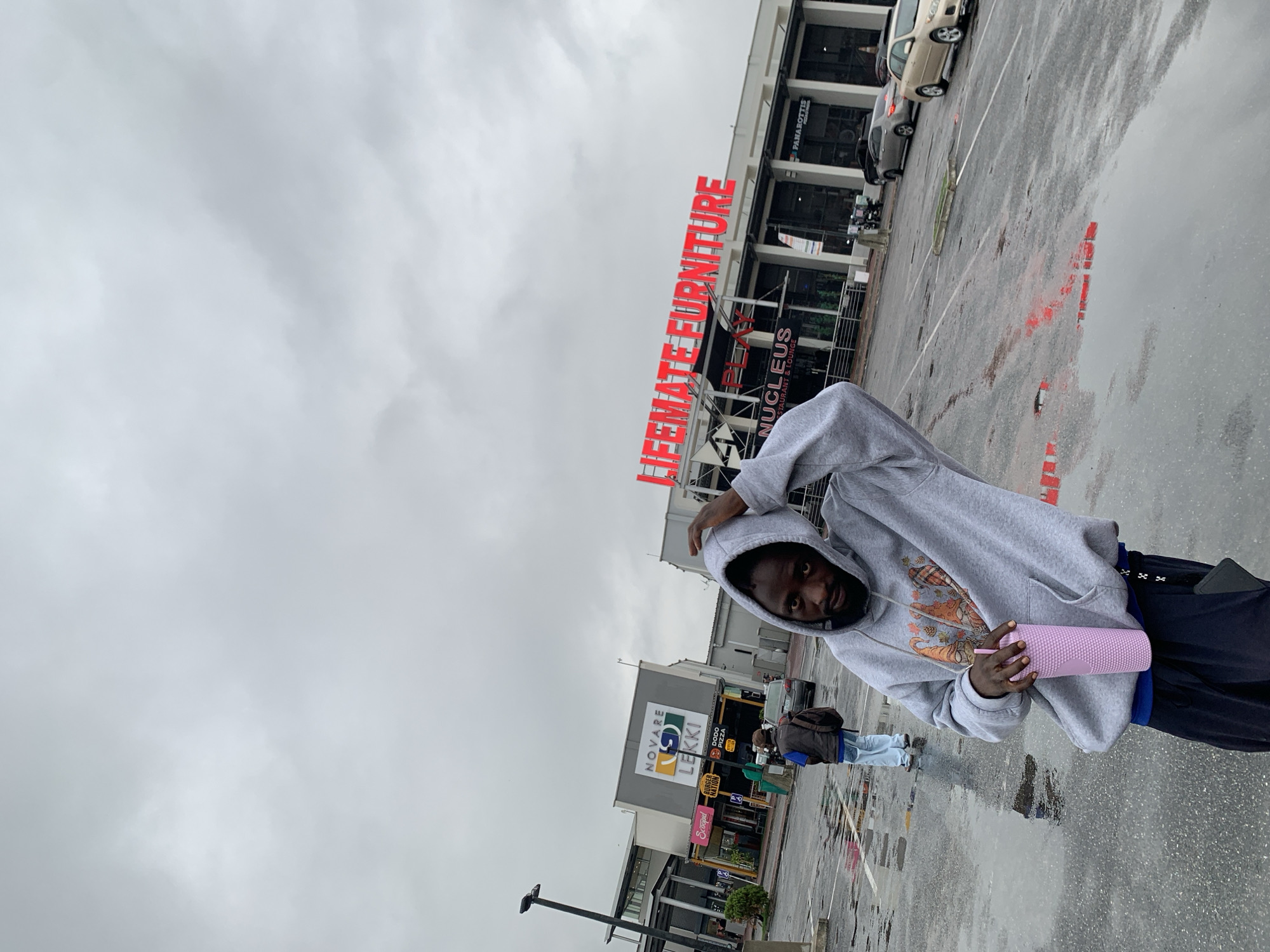 The image depicts a man standing in a parking lot in Sangotedo, Nigeria. The Lifemate Furniture store is visible in the background, along with the Novare Lekki mall sign. The ground is wet, indicating recent rainfall. The man is wearing a grey hoodie and holding a pink cup. He appears to be posing for the photo.