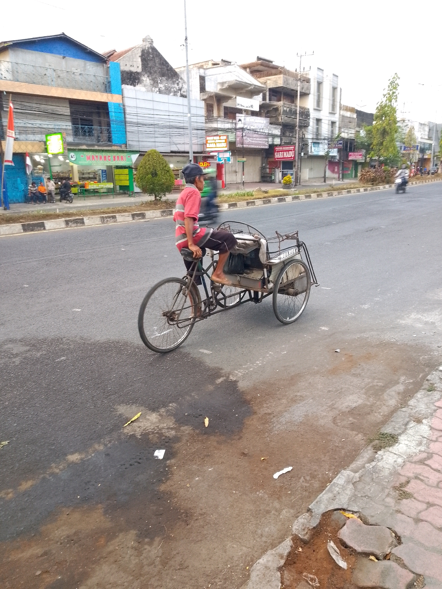 A person wearing a red and white striped shirt and dark shorts is riding a pedal-powered rickshaw on a paved road. The rickshaw has two wheels at the back and one at the front. The person is seated behind the handlebars, with their legs positioned as if pedaling. The rickshaw has a metal frame and a seat, with a covered compartment behind the rider. The road surface is asphalt with some wet patches and debris. On the left side of the road, there are buildings with various signs, including one with "MAYANG SARI" written on it. A red and white flag is also visible on the left. Several people are sitting on a bench in front of one of the buildings. In the background, there are more buildings and trees, suggesting an urban environment. A motorcycle is visible in the distance on the road. The sky is overcast.