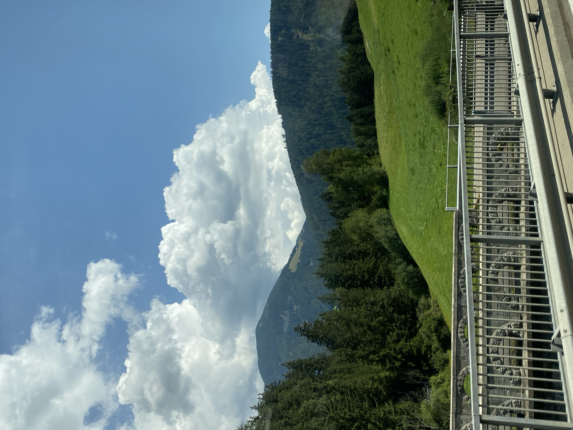 The image captures a scenic view of a mountainous landscape under a bright blue sky with scattered white cumulus clouds. In the foreground, a modern metal guardrail runs across the bottom of the frame, obscuring a concrete structure and a stone bridge with decorative arched details. Beyond the bridge, a lush green grassy hillside slopes upwards, dotted with scattered trees. Further in the background, dense evergreen forests cover the lower slopes of rolling mountains, which transition to a more sparsely vegetated, higher peak in the distance. The overall scene suggests a clear, sunny day in a rural, mountainous region. The location is identified as Mitterolang - Valdaora di Mezzo, Italy, which is consistent with the Alpine scenery depicted. There are no people or animals visible in the image, and no apparent actions or interactions taking place.