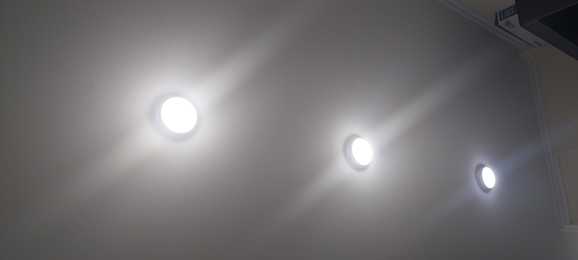 The image shows a ceiling with three recessed circular lights. The lights are illuminated, casting beams of light onto the textured ceiling. The lights are positioned diagonally from the bottom left to the top right of the frame. The ceiling appears to be white or off-white. There is a decorative molding visible along the bottom edge of the image, suggesting an indoor space. A partial view of a box with the Samsung logo is visible in the bottom right corner. The scene is indoors, and the location in the city of Unknown, Uzbekistan, is not determinable from the visual information provided.