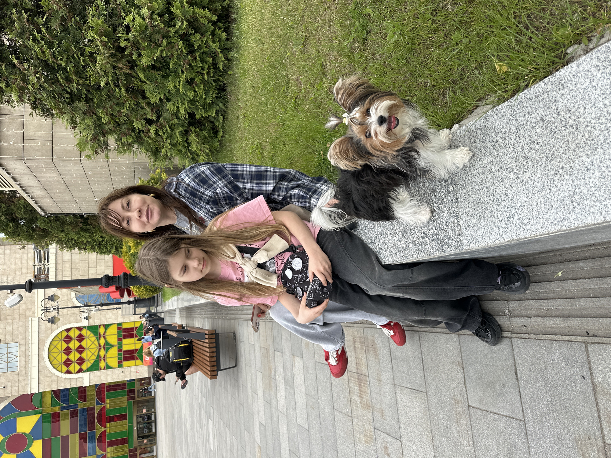 This outdoor image, taken in Moscow, Russia, captures two females and a small dog posing cheerfully on a low granite wall. The older female, with brown hair and bangs, wears a blue and white plaid shirt. Beside her, a younger girl with long brown hair wears a pink top, dark gray jeans, and red sneakers, holding a black bag adorned with white Hello Kitty patterns. A light-colored sweater is draped over her shoulders. The small tri-color dog, featuring black, white, and brown fur and a small bow in its topknot, sits attentively with its tongue slightly out, looking directly at the camera.
Behind the subjects, lush green grass and evergreen shrubs line the area. In the background, a large building features distinctive, colorful stained-glass-like windows with geometric red, yellow, green, and blue patterns. A paved pedestrian walkway with square tiles extends into the foreground, where several lampposts and public benches are visible, some occupied by other people relaxing. The scene suggests a pleasant daytime, likely during late morning or afternoon, with clear weather. The subjects appear relaxed and happy, enjoying a public space.