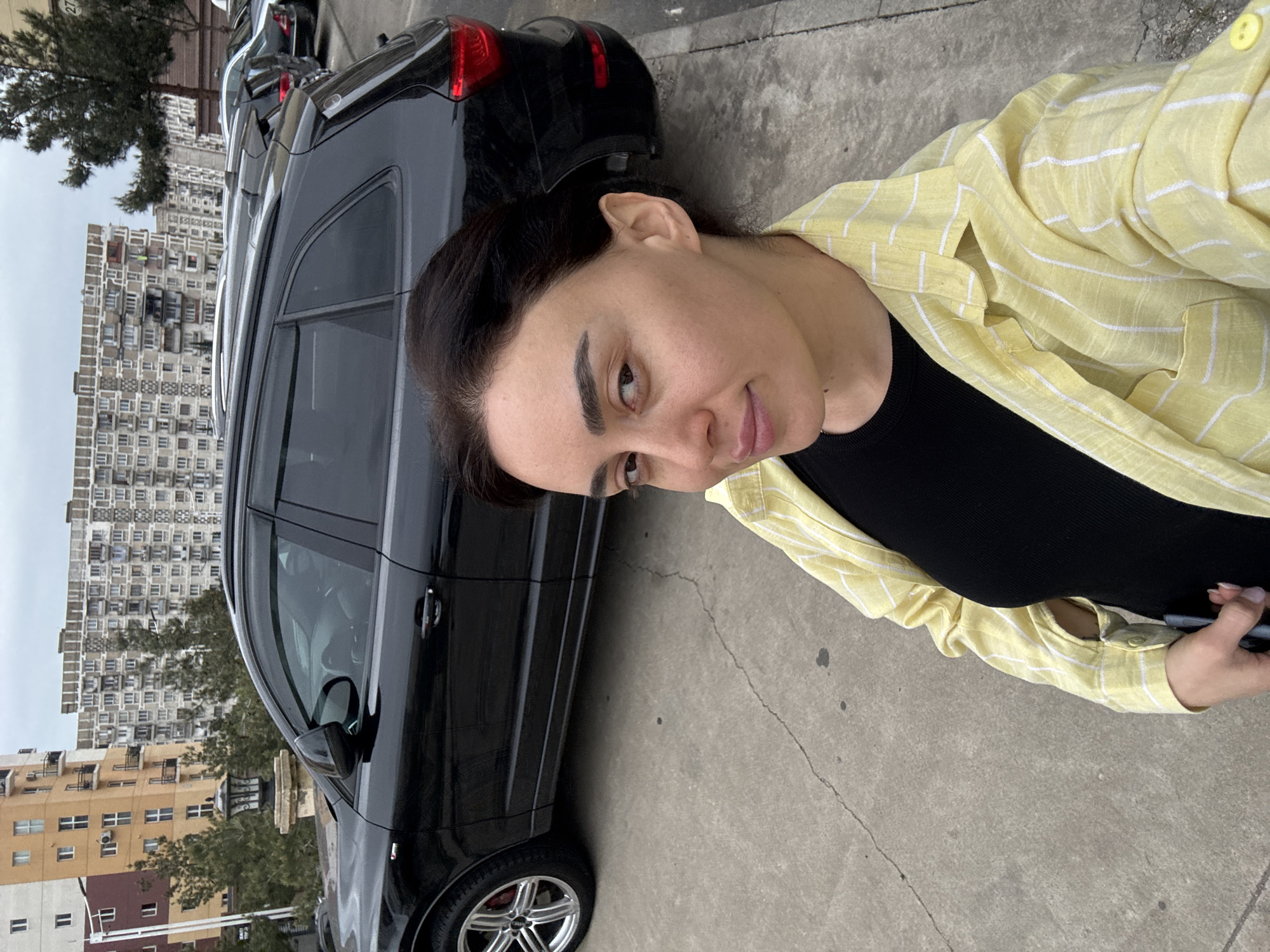 The image captures a woman taking a self-portrait outdoors in an urban environment under an overcast sky. The woman is in the foreground, looking directly at the camera with a neutral expression and a slight smile. She has dark hair, dark eyebrows, and is wearing a black top underneath a yellow and white striped long-sleeved button-down shirt. Her right hand, holding a smartphone, is visible at the bottom right of the frame.
Behind the woman, a black SUV is parked on a concrete surface, with its side visible. The vehicle has silver multi-spoke wheels and roof rails. In the background, several multi-story apartment buildings are visible. One building particularly stands out with numerous small, uniform windows or balconies, characteristic of Soviet-era residential architecture. Trees are scattered between the buildings and the parked car. The overall setting appears to be a street or parking area in a city, identified as Tbilisi, Georgia. The weather is overcast. No visible text is legible in the image.