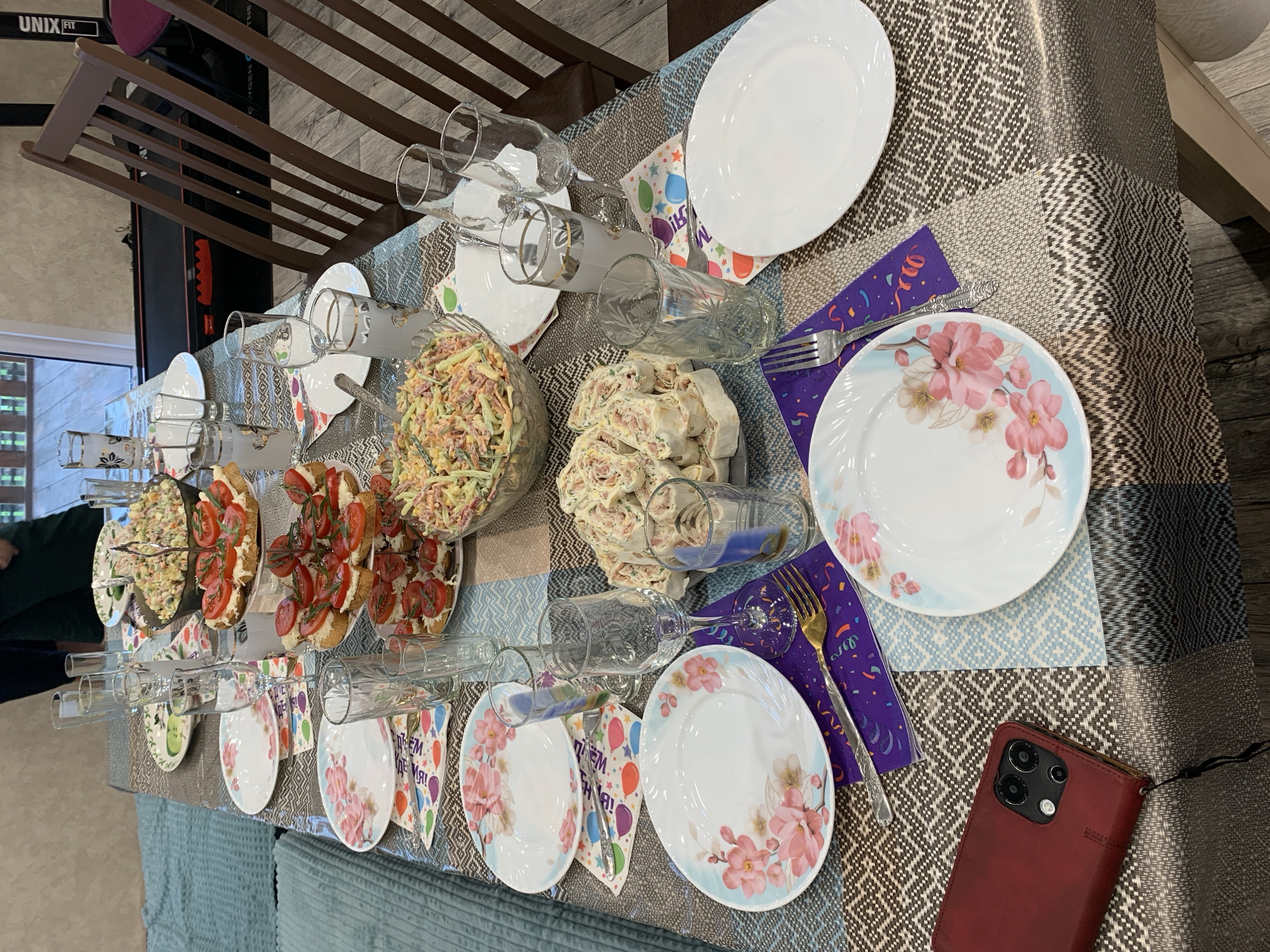 The image depicts a close-up, eye-level view of a table set for a meal, likely in a home environment. The table is covered with a patterned tablecloth. On the table are several plates, some floral-patterned and others plain white. There are also various glasses, some containing a light-colored liquid, and silverware, including forks and what appears to be a serving spoon in a bowl of salad.
The food items visible include a large salad in a clear bowl, what looks like canapés or small sandwiches topped with tomatoes, and rolled wraps, possibly filled with a creamy mixture and herbs. Several patterned napkins, some with confetti and celebratory motifs, are placed under the plates and cutlery.
In the background, part of a wooden chair with dark brown slats is visible, along with a treadmill and a portion of a wall. A red smartphone in a wallet-style case rests on the tablecloth in the lower left corner. The overall impression is one of a prepared gathering or celebration. The provided image does not contain any information to suggest the location is in the city of Asha, Russia.