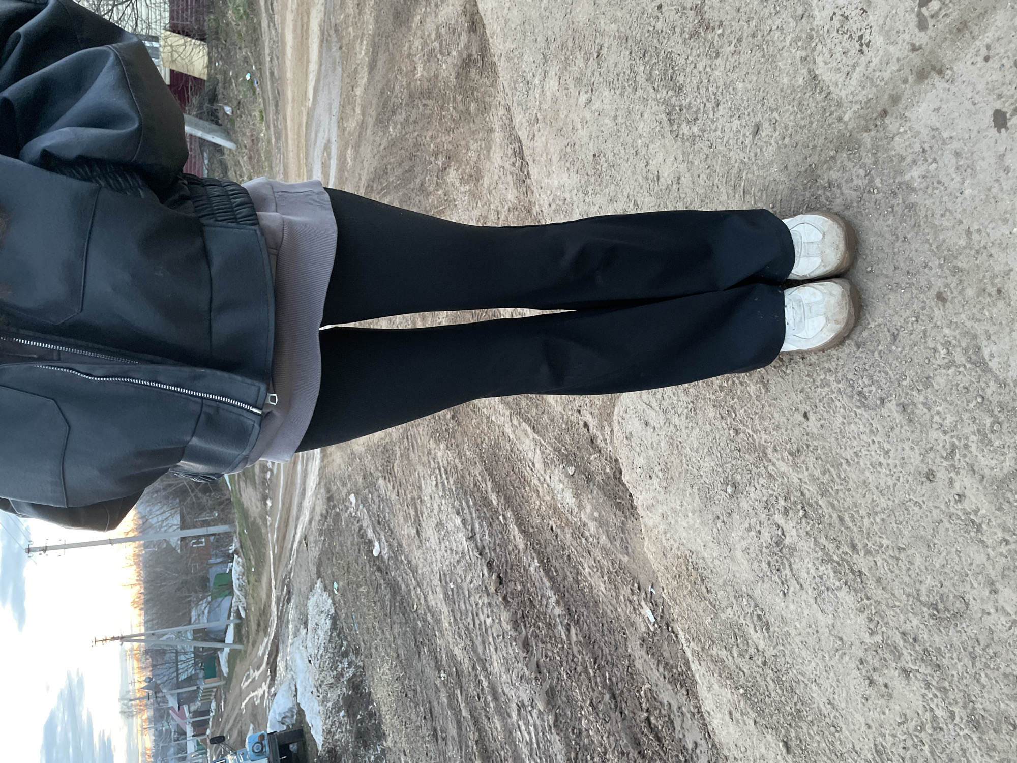 The image shows a person standing on a dirt road in Фоки, Russia. The person is wearing a black leather jacket, a grey sweatshirt, black pants, and dirty white shoes. The road is uneven and muddy, with evidence of tire tracks. In the background, there are houses, trees, and telephone poles. The sky is visible in the distance, with some clouds present. It appears to be a cool or cold day.