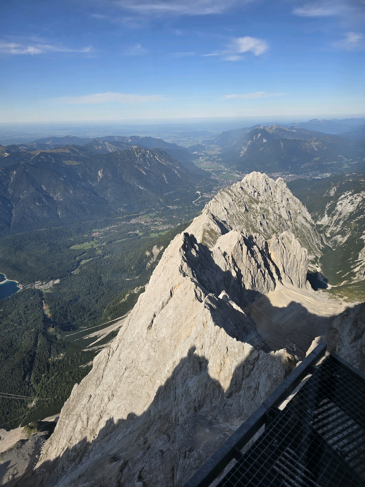 The image is a high-angle, telephoto view of the Zugspitze mountain range, specifically looking out over the valley from a peak in Grainau, Germany. The foreground features a metal grate platform, casting a shadow on the rugged, rocky mountainside. The mountains are sharp and jagged, with visible striations in the rock.
In the middle ground, a sharp, prominent peak dominates the scene, with a valley floor visible behind it. The valley is covered in dense, dark green evergreen forests. A small, turquoise lake is visible on the left side of the valley. A cable car with red and orange carriages is suspended in the air, with cables extending down towards the lake and the forest. A small town or village is nestled within the valley, with buildings appearing as small clusters of lights and structures.
The background shows a series of layered mountain ranges fading into a hazy horizon. The sky is a clear, bright blue with a few wispy white clouds. The sun appears to be casting long shadows, suggesting it is either early morning or late afternoon. The overall impression is one of vastness and natural beauty, with human infrastructure, like the cable car, integrated into the landscape.