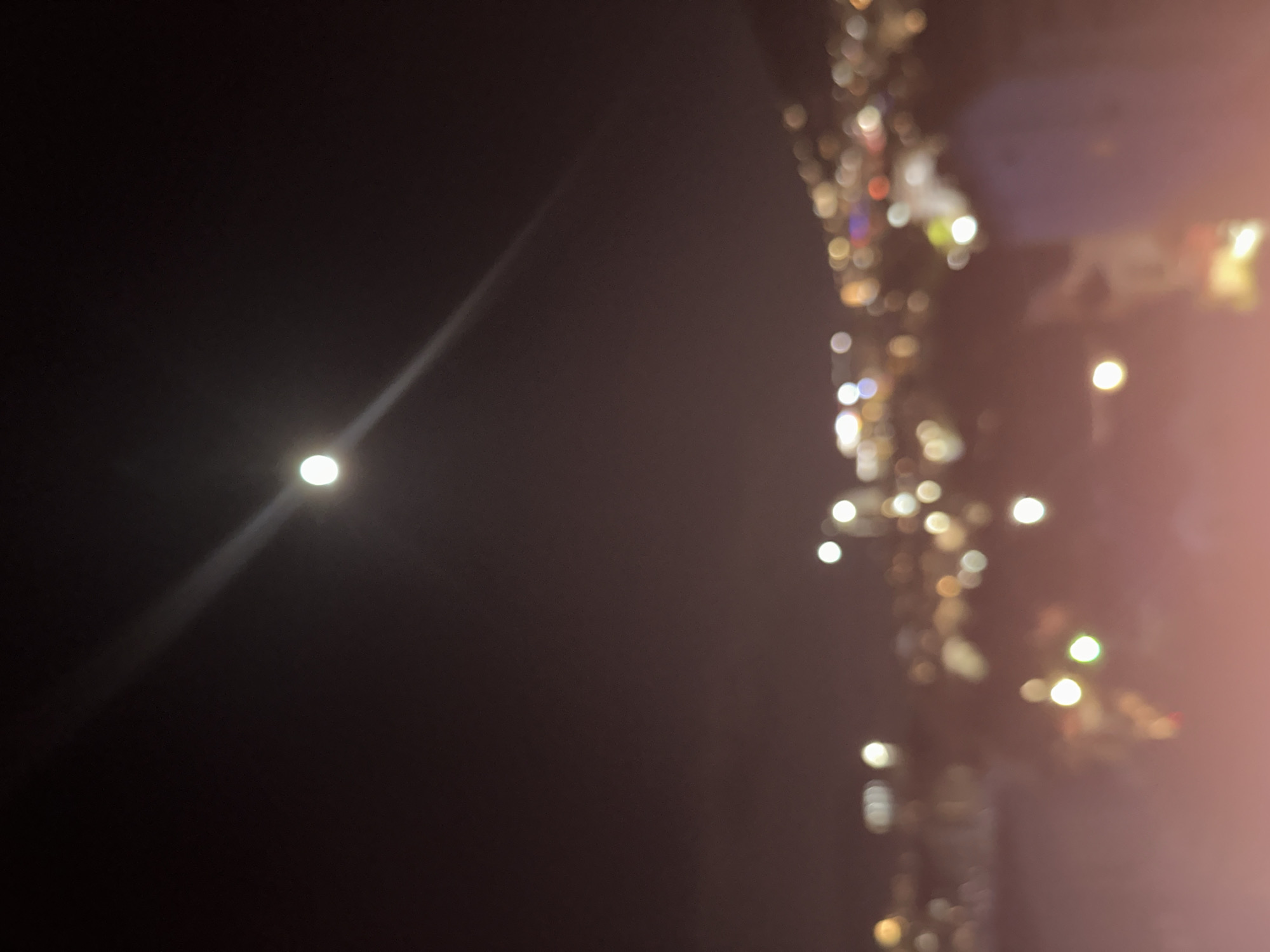 Nighttime image shows city lights on the right of the frame, blurred and out of focus. On the left is a bright light source, also blurred, with a streak extending from it. The overall color palette is dark, with the blurred lights providing accents of yellow, white, and hints of red and blue. It is not possible to identify the exact location in the city of Budva from the given image.