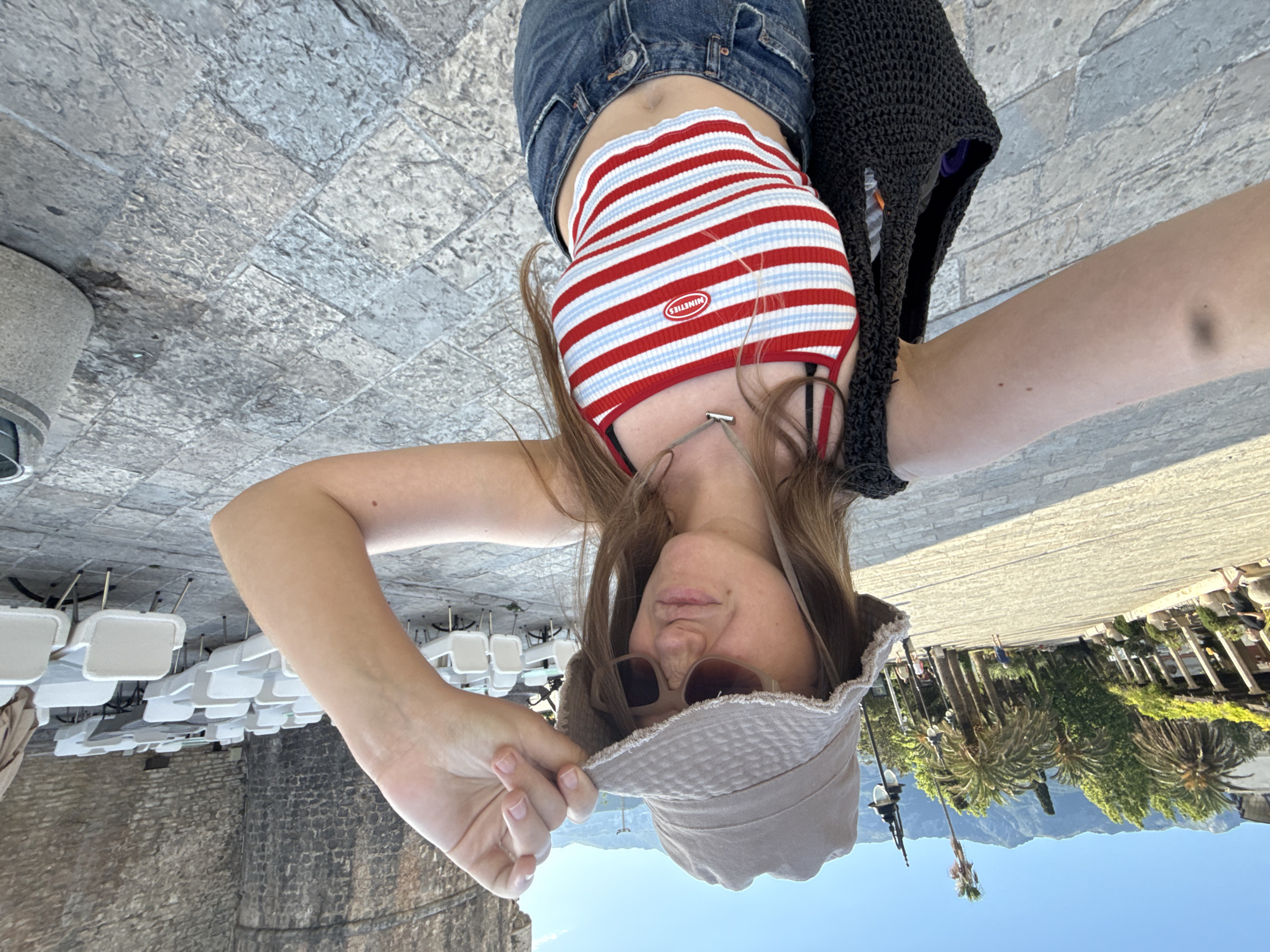 An outdoor selfie captures a young woman in Budva, Montenegro, on a sunny day. The subject, a female with long brown hair, occupies the foreground, looking directly at the camera with a slight puckered expression and wearing dark sunglasses and a beige, distressed bucket hat. Her left hand adjusts the hat brim. She wears a red, white, and light blue striped crop top with an oval "NINETIES" emblem on the chest, and blue denim jeans. A black woven bag is slung over her right arm, and a small, dark mark is visible on her upper right arm.
The environment behind her is characterized by ancient stone architecture and paved ground. To her right, a tall, rough-hewn stone wall dominates the background, likely part of a historic structure. In front of this wall, a large, organized arrangement of empty white chairs and matching tables indicates an outdoor dining area. To her left, beyond a paved square, a pedestrian path is visible, lined with palm trees and other green foliage. A wooden pergola structure with climbing plants shades a section of the path, under which a person walks. Distant mountains rise under a clear blue sky. The ground is paved with irregular grey and tan stone blocks. A stone or concrete trash receptacle is partially visible in the lower right corner.