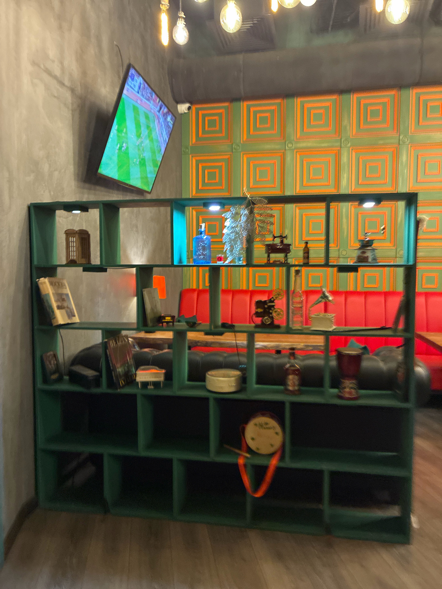 The image depicts the interior of a modern establishment, likely a bar or restaurant, with a distinctly contemporary and eclectic design. A large, dark gray concrete wall dominates the left side of the frame, with a flat-screen television mounted on it, displaying a soccer game. Above the TV, exposed filament light bulbs hang from the ceiling, contributing to the industrial-chic aesthetic.
The central focus of the image is a tall, dark green shelving unit filled with various decorative objects. These include a miniature red British telephone booth, books, bottles of liquor, a vintage sewing machine, a projector, a gramophone, and a small drum with an orange ribbon. The shelves are illuminated by small, embedded lights.
Behind the shelving unit, a vibrant wall features a repeating pattern of green and orange squares. A long, bright red booth seating area runs along the back wall, suggesting a comfortable dining or lounging space. The floor is made of light-colored wooden planks.
The overall impression is a well-designed space that combines industrial elements with decorative touches, creating a visually interesting and possibly lively atmosphere. The presence of the television displaying a sports game and the bar-like seating indicates a venue geared towards entertainment and socializing. The specific location in Sochi, Russia, is not discernible from the visual elements alone.