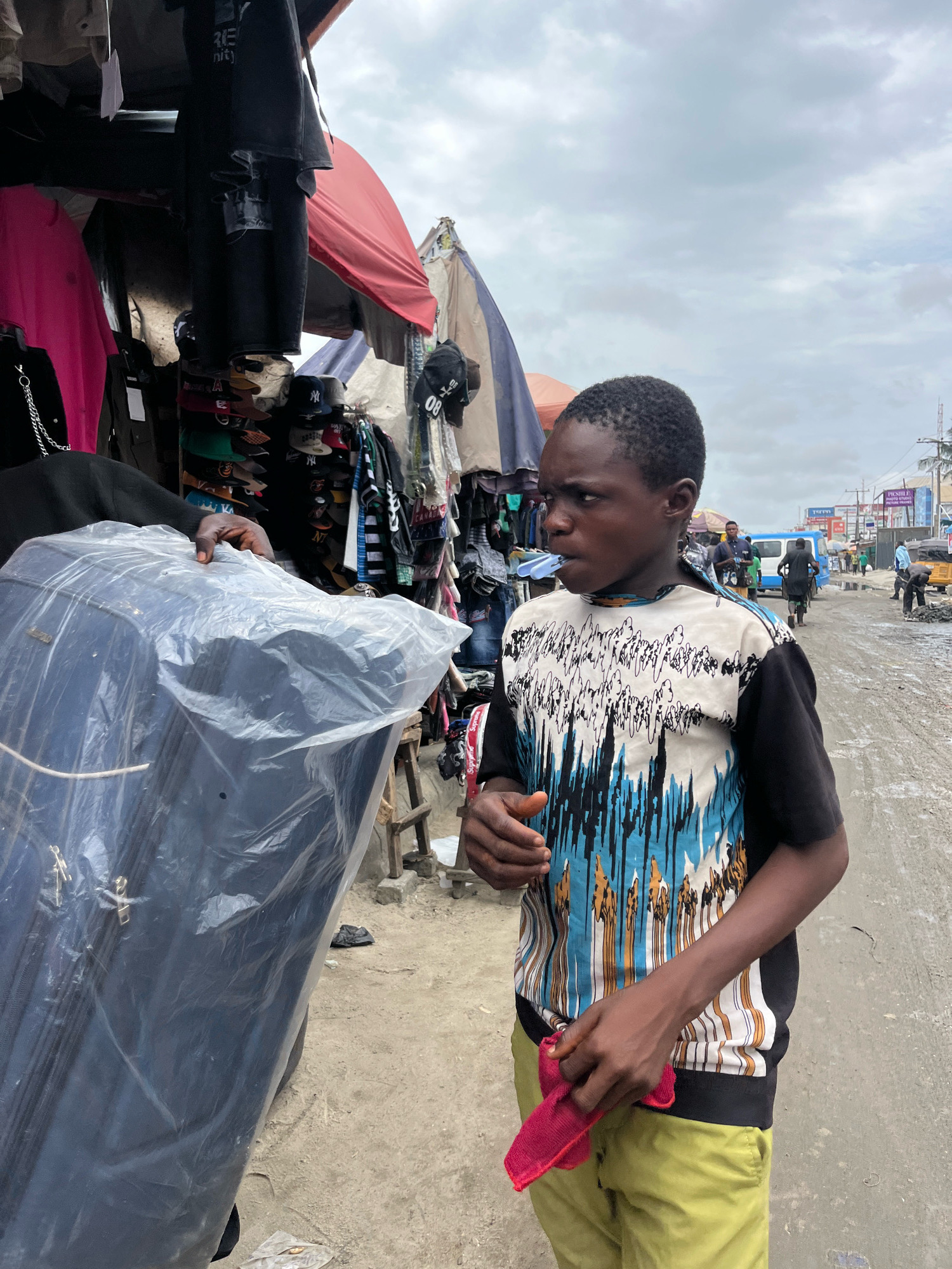 A young boy, appearing to be in his early teens, stands in a bustling outdoor market in Ajah, Nigeria. He is wearing a black and white patterned t-shirt with blue and brown abstract designs, and light olive-green pants. In his right hand, he holds a small, folded red cloth. His left arm is partially obscured by a large, transparent plastic-wrapped object, possibly a mattress or large piece of fabric, which he is holding with another person whose arm is visible.
The boy is looking to his right, with a focused expression. Behind him, a market stall is laden with various clothing items, including t-shirts, pants, and hats, some of which are hanging from racks. The ground appears to be a dirt and concrete surface, typical of a market area. In the background, other people are visible, along with motorbikes, a blue bus, and the general activity of a roadside market. The sky is overcast, suggesting a cloudy day. The overall scene depicts a lively commercial environment.