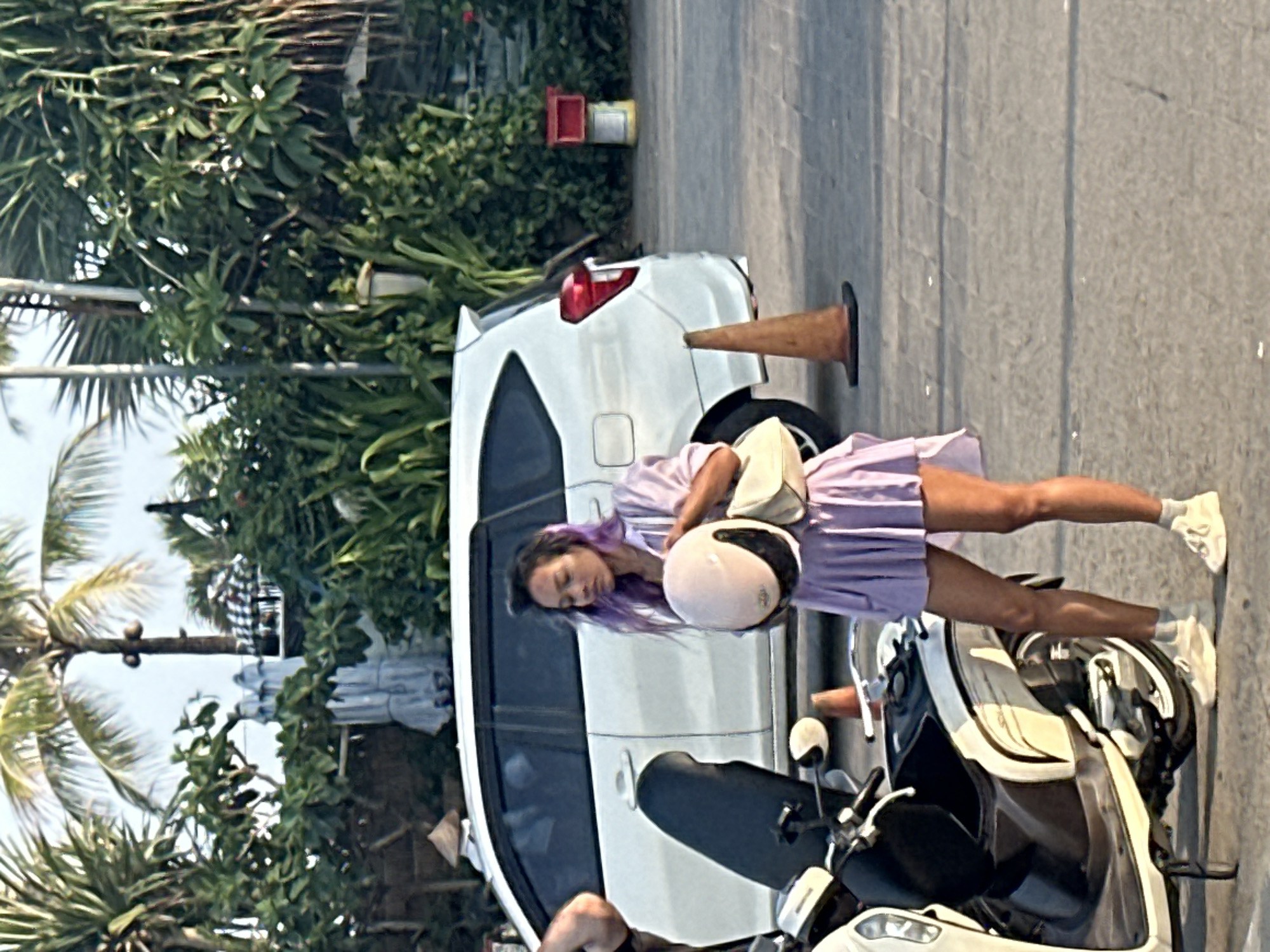A woman with purple-dyed hair stands next to a white scooter in North Kuta, Indonesia. She is wearing a light purple, pleated dress and holds a light pink helmet and a cream-colored handbag. A white car is parked behind her, and a traffic cone is visible on the right side of the image. The background consists of lush green foliage and palm trees. The ground is paved with stone tiles. The scene appears to be an outdoor area with a tropical climate.