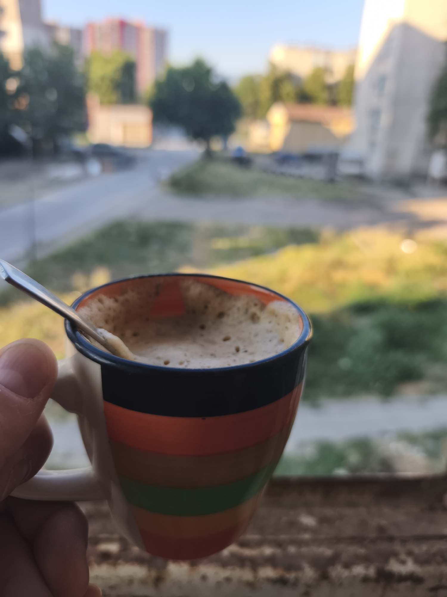 An outdoor medium shot shows a hand holding a colorful striped mug filled with a frothy beverage, likely coffee, with a spoon resting inside. The mug has horizontal stripes of orange, black, green, and red. The background is blurred, depicting an urban environment with buildings, trees, and a road. The lighting suggests it is either early morning or late afternoon, with the sun casting a warm glow on the scene. The overall impression is a moment of quiet enjoyment in an everyday urban setting.