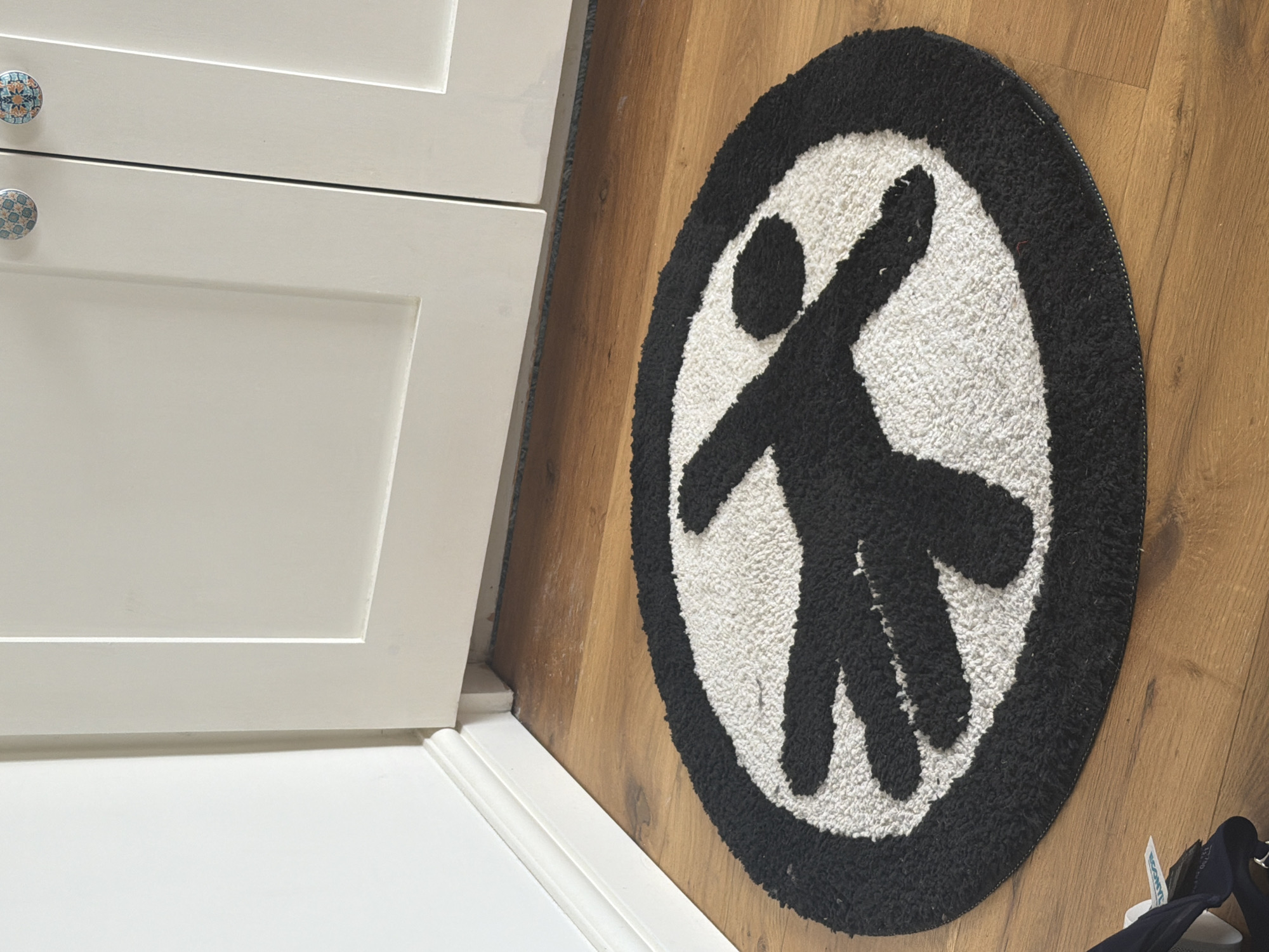 The scene, located in the city of London, United Kingdom, depicts an indoor environment. A circular rug, approximately 0.7 meters in diameter, lies on a light brown wooden floor. The rug features a black, stylized silhouette of a human figure with outstretched limbs against a textured white background, all contained within a black circular border. The wooden floor exhibits visible grain patterns. In the background, a white wall with a white skirting board is visible on the left. Centrally and to the right, two off-white, paneled cabinet doors are present, each equipped with a decorative, patterned circular knob. A dark, recessed area is visible beneath the cabinet base, directly above the floor. No individuals, active subjects, or interactions are depicted.