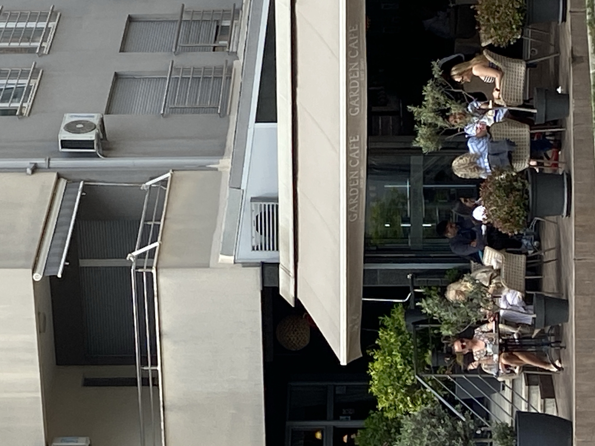 The image depicts an outdoor patio area of "Garden Cafe," presumably in the city of Budva, Montenegro. Several individuals are seated at tables. There are four people at one table: one woman with blonde hair in a striped shirt and sunglasses. There are two people at another table: one woman with blonde hair and a white shirt and one man wearing a dark top. The third table has three people: one woman with blonde hair and two people wearing dark tops. The patio is sheltered by a white awning, with the cafe's name emblazoned on its side. Foliage and potted plants are interspersed between the tables. An adjacent building has a grey facade with air conditioning units and windows with metal bars.