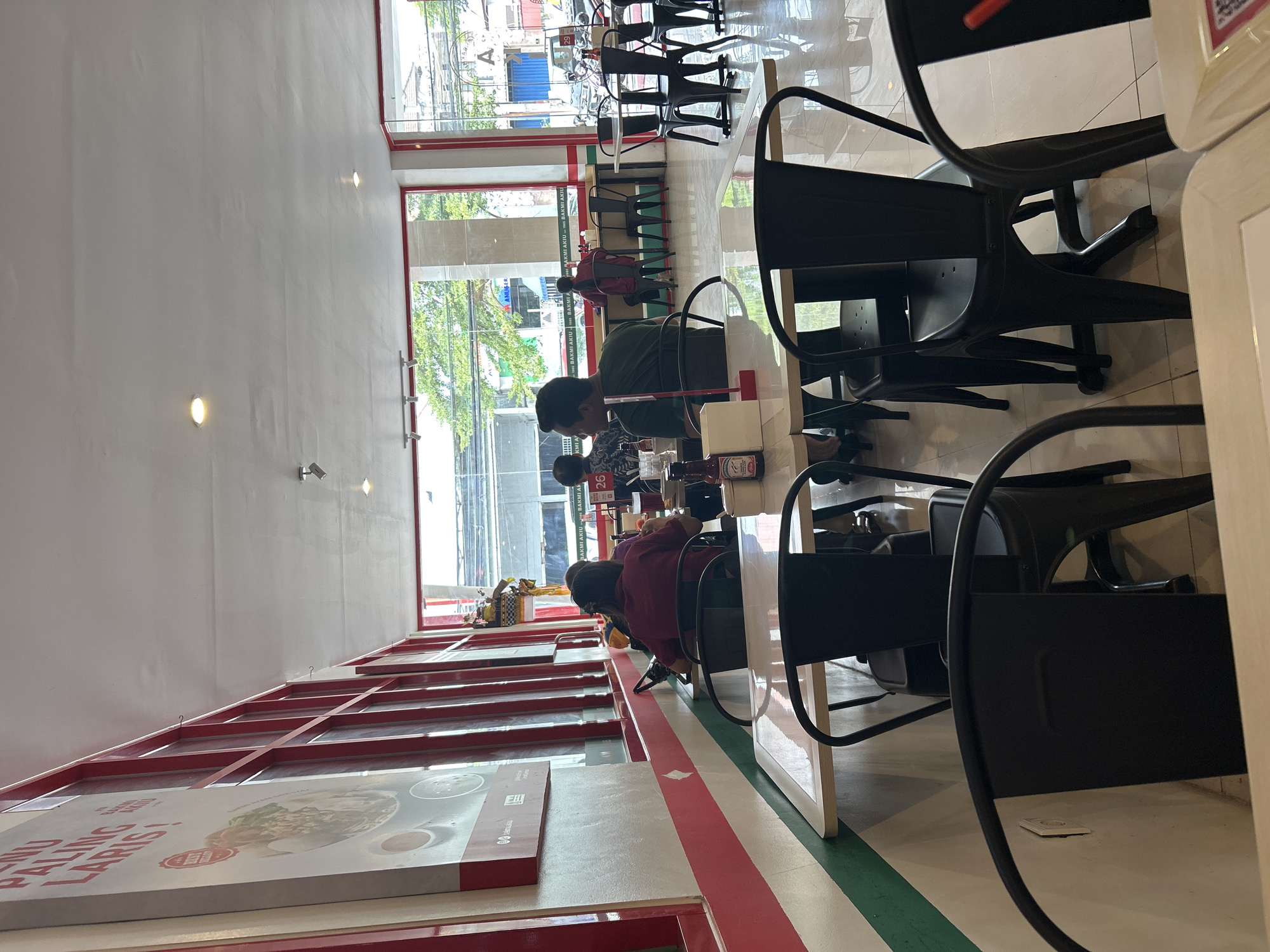 This is an interior shot of a restaurant in Denpasar, Indonesia. The restaurant features a bright, modern design with white walls, red accents, and a tiled floor. Several tables and chairs are visible, some occupied by customers. A large poster with Indonesian text, likely advertising menu items, is mounted on the wall. Through the large windows at the back of the restaurant, a street scene with trees and buildings can be seen. The atmosphere appears casual and busy.