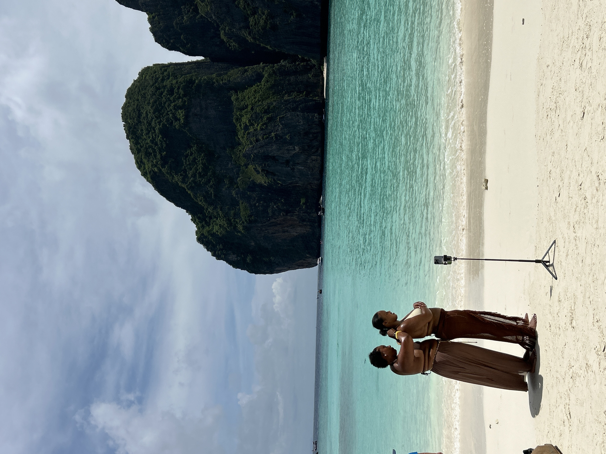 Two women are standing on a sandy beach in Ao Nang, Thailand, with turquoise water and large limestone cliffs in the background. The woman on the left, wearing a brown skirt, has her arms around the woman on the right, who is wearing a brown sarong. A smartphone on a tripod is positioned in front of them, suggesting they are either taking a photo or recording a video. The sky is partly cloudy. The ocean is calm, with small waves breaking on the shore. Several boats are visible in the distance. The overall scene depicts a tropical beach setting.