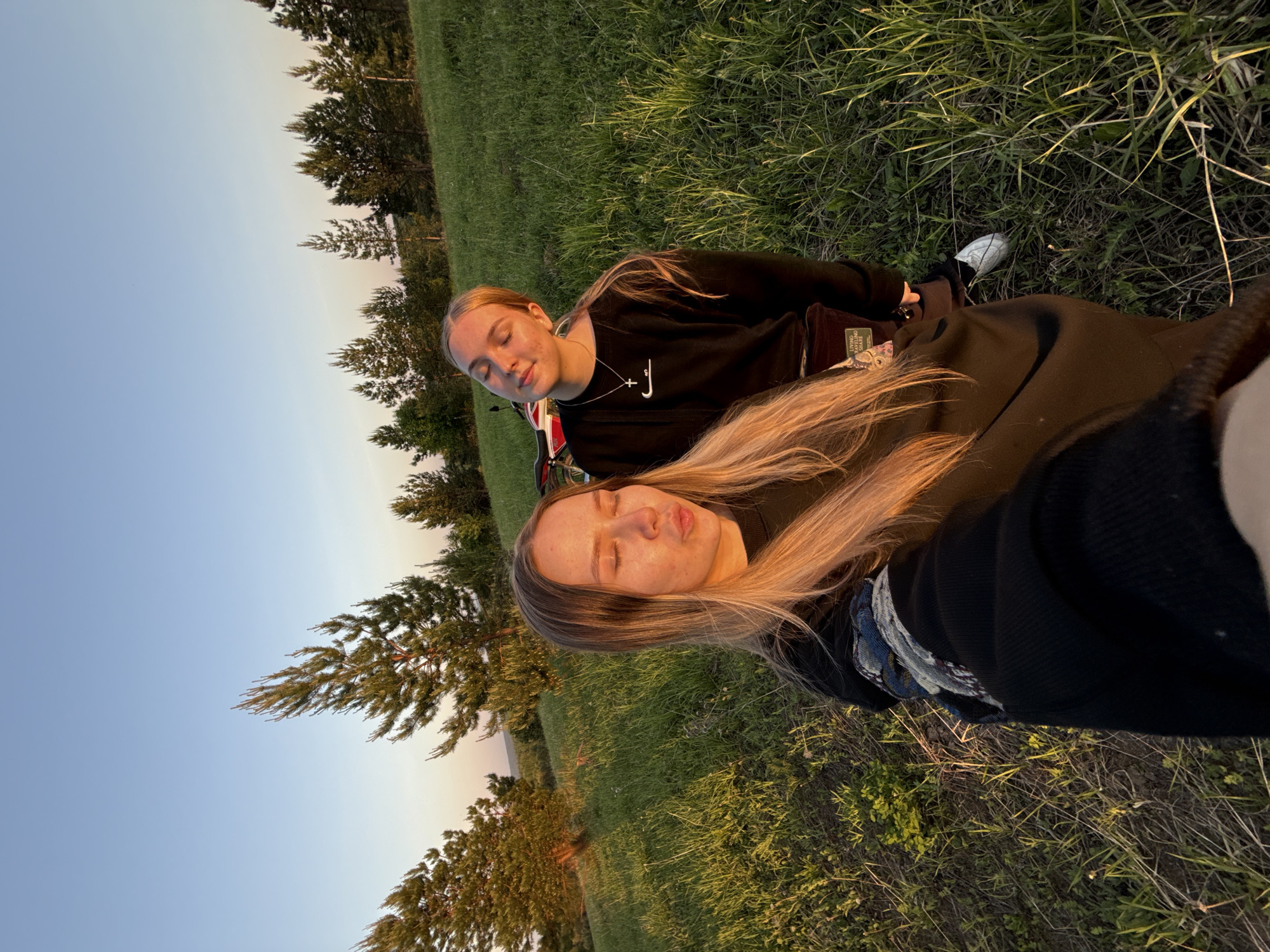 Two young women are lying on a grassy field at dusk. The woman in the foreground has blonde hair and is wearing a black sweatshirt. The woman behind her has similar hair and is also wearing a black sweatshirt with a Nike logo. Trees are visible in the background. The location is likely Zhigalki, Russia.