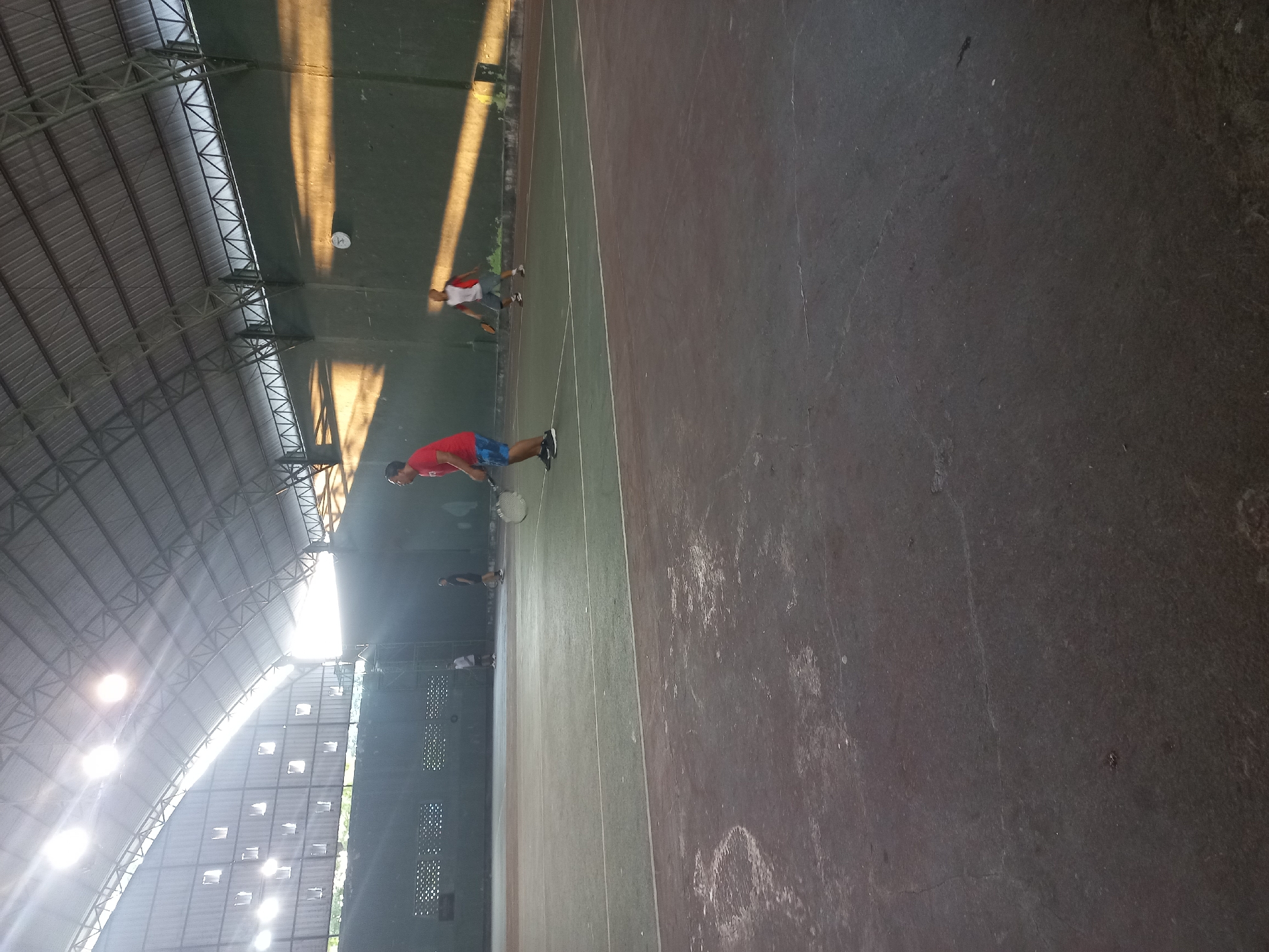 The image depicts an indoor tennis court in Danyang, Indonesia. The court has a dark grey surface with lighter grey markings indicating boundaries. Several people are present on the court, engaged in playing tennis. One individual in the foreground on the right is wearing a red shirt and blue shorts, holding a tennis racket and appearing to be mid-action. Another person in the background on the left is wearing a white shirt and dark shorts, also with a racket. Two other individuals are visible further in the background, one in a dark shirt and the other in a white and red shirt. The court is covered by a large, grey, curved roof supported by a metal frame. Natural light streams in from openings in the roof structure, creating distinct sunbeams that illuminate parts of the court and walls. The walls of the enclosed space are dark green. The overall impression is of an active sports facility during daylight hours.