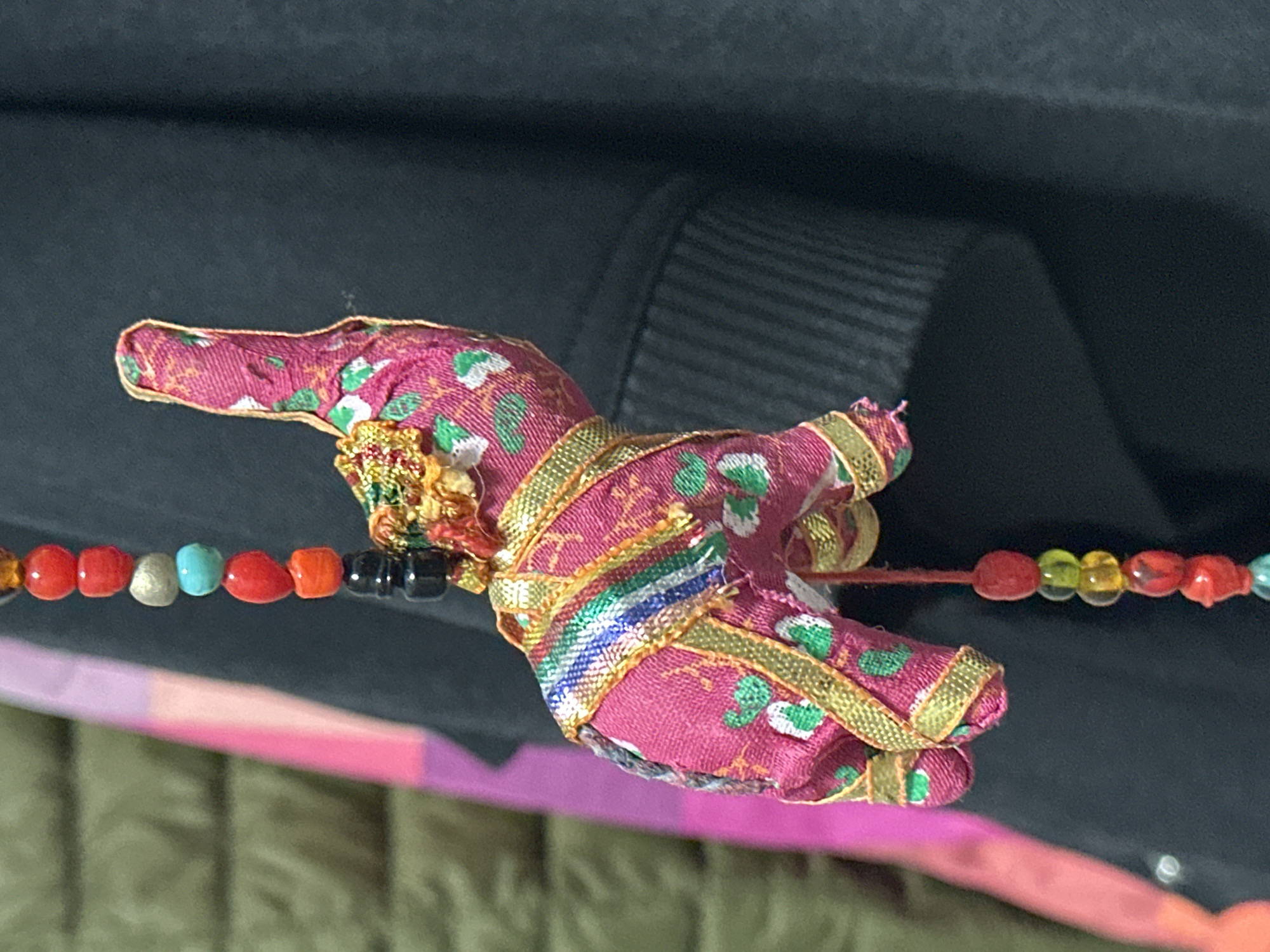 The image is a close-up, eye-level shot of a handmade fabric elephant charm hanging from a beaded string. The elephant is primarily pink with a pattern of green dots and white flowers. It is adorned with gold trim and a decorative, golden headpiece. The beaded string above the elephant consists of various colored beads, including red, orange, black, turquoise, and grey. Below the elephant, a similar beaded string with red, yellow, and light blue beads is visible. The background is dark and out of focus, showing hints of a textured surface that could be fabric or clothing. The lighting is uneven, casting shadows. The scene is devoid of any identifiable landmarks or characteristics that would place it specifically in the city of Nevinnomyssk, Russia.