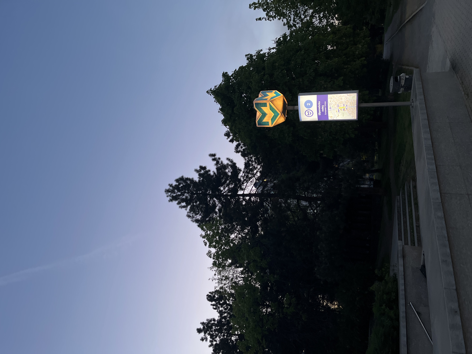 The image depicts an outdoor scene at what appears to be a metro station entrance, viewed in portrait orientation as if the camera was rotated 90 degrees clockwise. In the center, an illuminated, three-dimensional metro sign stands prominently. The sign is shaped like a blocky letter "M" with arrows pointing towards its center, glowing yellow and green.
To the right of this sign, an illuminated rectangular information panel is mounted on a pole. The panel displays a large circled number "2" and a metro symbol (a stylized "M"). Below these, the text "Европейски съюз" in Cyrillic is visible, followed by its English translation "European Union." A simplified metro map with colored lines and station names is also displayed on the right side of this panel.
The background is dominated by lush, dark green trees and foliage, suggesting a natural or park-like setting around the station entrance. Above the trees, the sky is a deep, clear blue, indicating either dusk or dawn. The illumination of the signs and the dim ambient light support this assessment. In the foreground and extending to the right, a concrete pathway or set of stairs with a short concrete wall is visible, leading downwards, presumably to the metro entrance. The overall scene suggests a quiet, early evening or morning atmosphere.