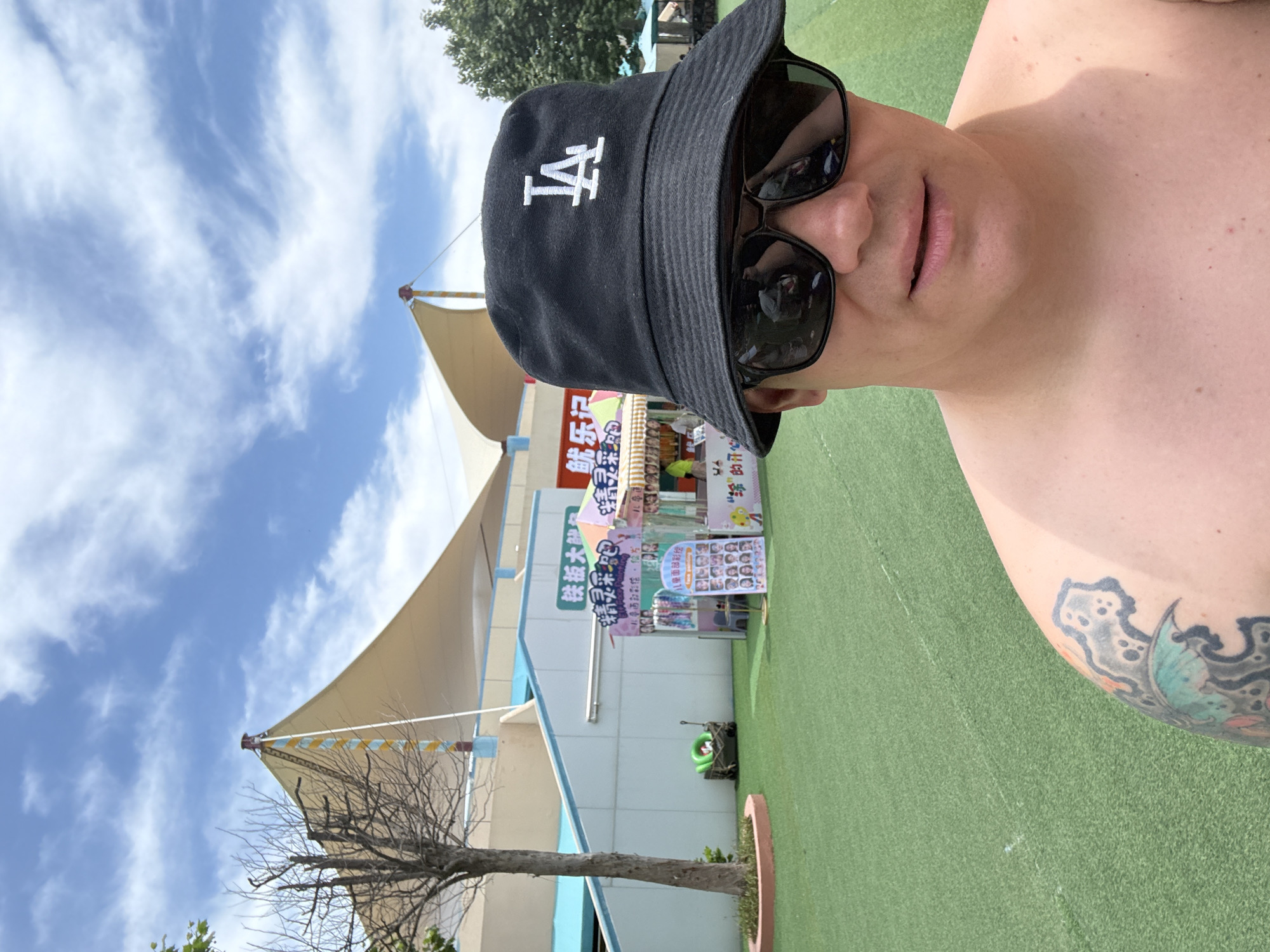 The image shows a selfie taken by a bare-chested man wearing a black bucket hat with an "LA" logo and black sunglasses. He has a tattoo on his left shoulder and upper arm. He is standing outdoors on a bright, sunny day. In the background, there is a large, beige, tent-like structure supported by poles, a building with blue and white trim, and some trees. To the left of the man, there is a small, bare tree. In the mid-ground, there are two vendor stalls set up on a bright green, artificial turf surface. One stall has a sign with Chinese characters and images of faces, suggesting it is a face painting booth. The other stall also has Chinese signage and colorful awnings. The overall setting appears to be an outdoor recreational area or amusement park.
This image was taken at the Nandaihe Tourist Resort in Qinhuangdao, China. The Nandaihe Tourist Resort is known for its beaches, amusement parks, and family-friendly attractions. The specific location appears to be within a section of the resort featuring outdoor entertainment and vendor activities, possibly near a play area for children. The structures in the background are characteristic of the resort's architectural style, designed to provide shade and a visually appealing environment for visitors. The vendor stalls are typical of those found in tourist areas, offering services like face painting and selling snacks or souvenirs.