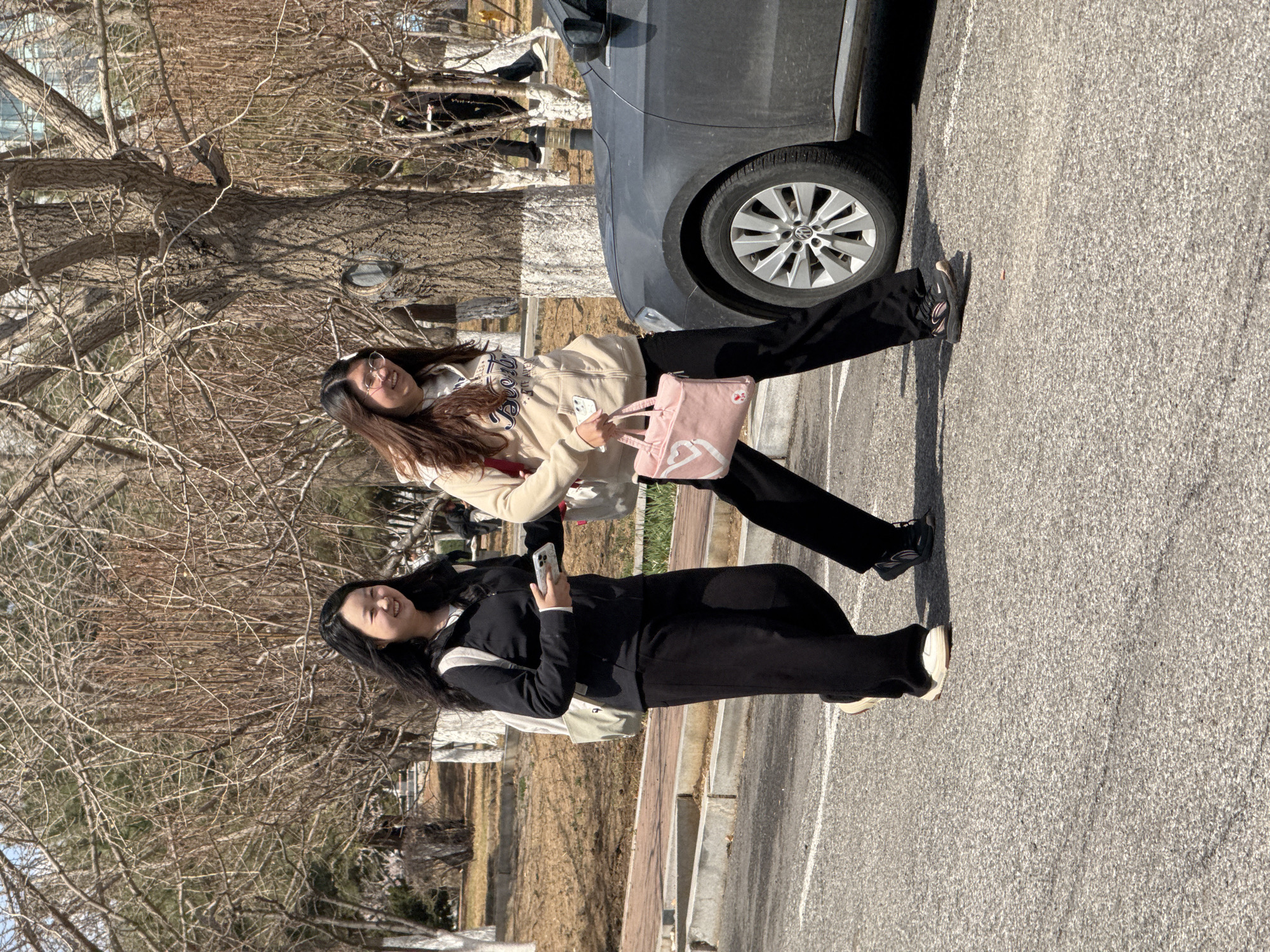 Two young women are standing next to a car in what appears to be a parking lot. The woman on the right is wearing a light-colored hooded jacket, black pants, and carrying a pink tote bag. The woman on the left is wearing a black jacket and pants. She is holding a smartphone. The background includes bare trees, some buildings, and a grassy area. The location is Zhongtun, China.