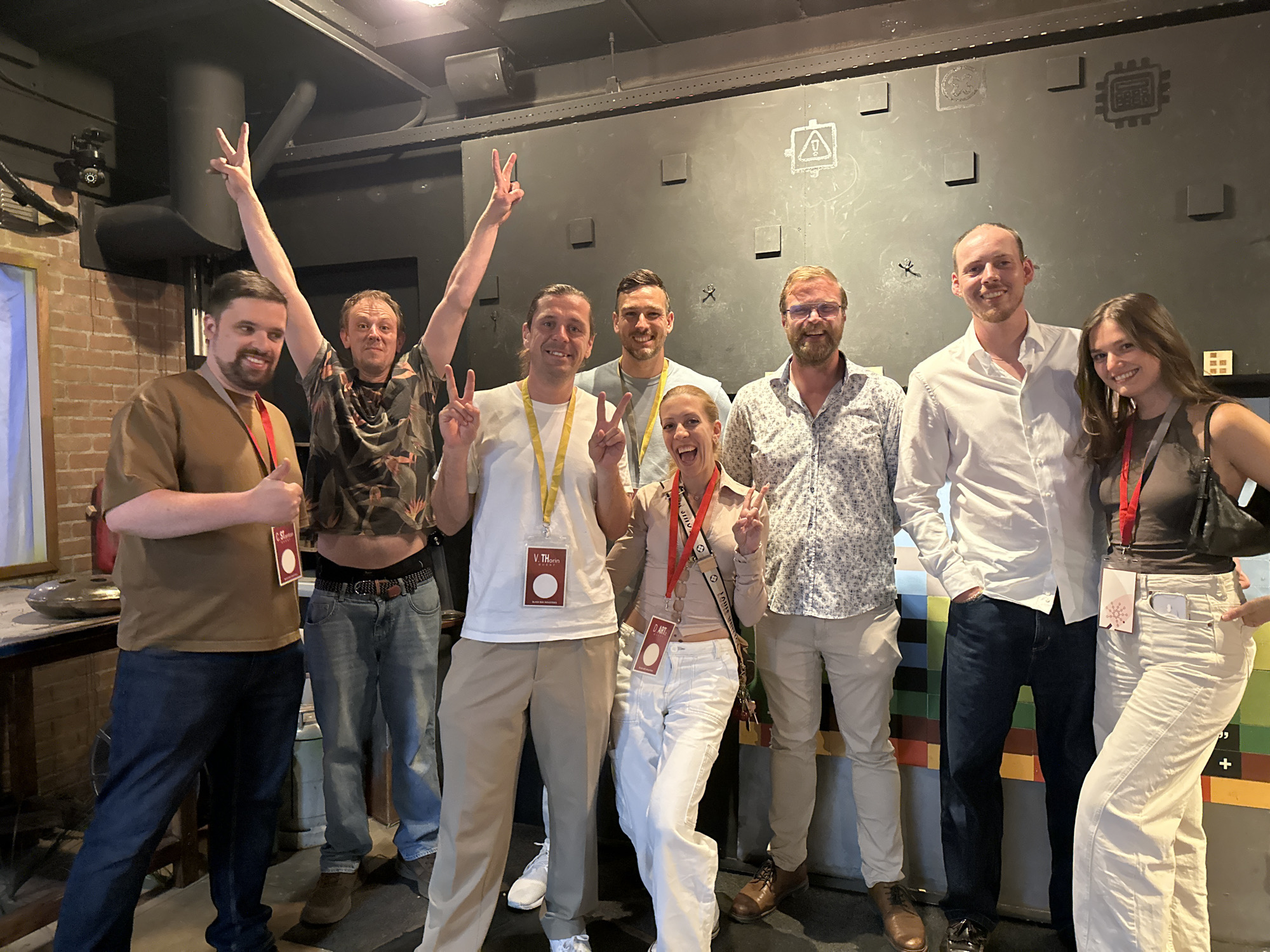 Eight people, seven men and one woman, are posing for a photo in an indoor setting. The background appears to be a dark, textured wall with some square or rectangular protrusions and stenciled symbols. The lighting is dim, suggesting an interior space like a venue or bar. The group is standing in front of this wall, with some individuals holding up peace signs with their fingers. The people are dressed casually, mostly in t-shirts, button-down shirts, and jeans. Several individuals are wearing lanyards with badges or passes around their necks, suggesting they may be at an event or conference. The overall atmosphere is relaxed and friendly, with most of the individuals smiling. No specific Amsterdam landmarks are visible in the image.