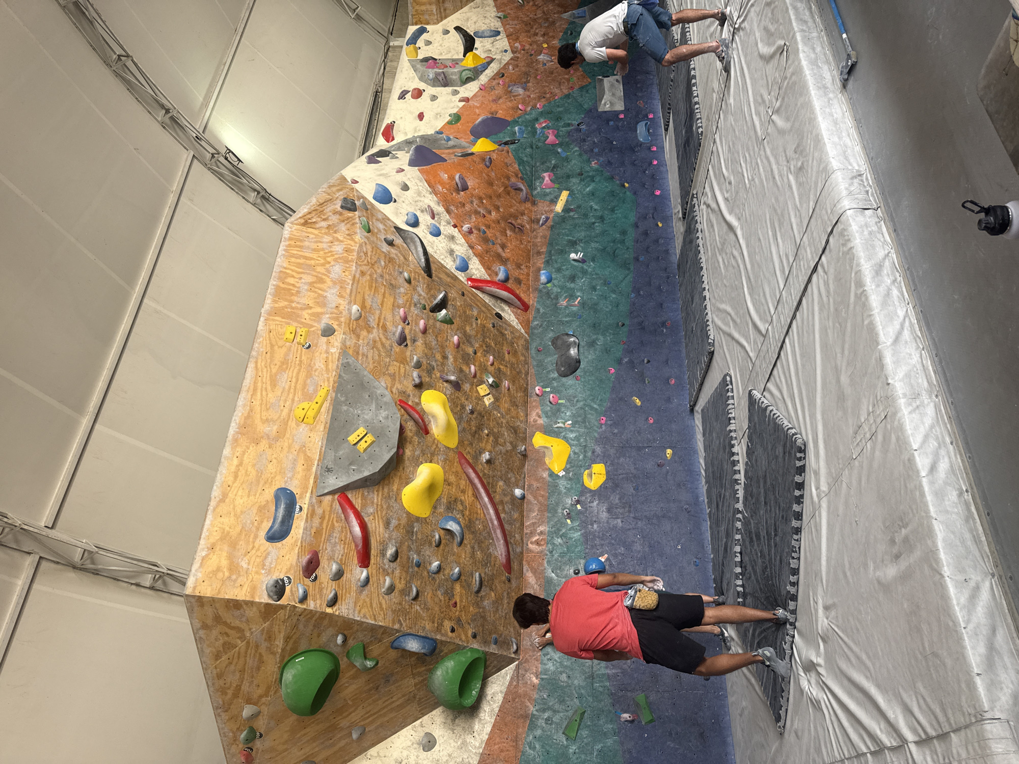 A climbing gym in San Andrés Cholula, Mexico shows two people climbing an indoor rock wall. The wall is multi-colored with various climbing holds attached to different sections. One climber wears a red shirt and black shorts, while the other wears a white shirt and blue shorts. They are positioned on different parts of the wall. The walls of the gym are white and the floor is covered with padded mats.