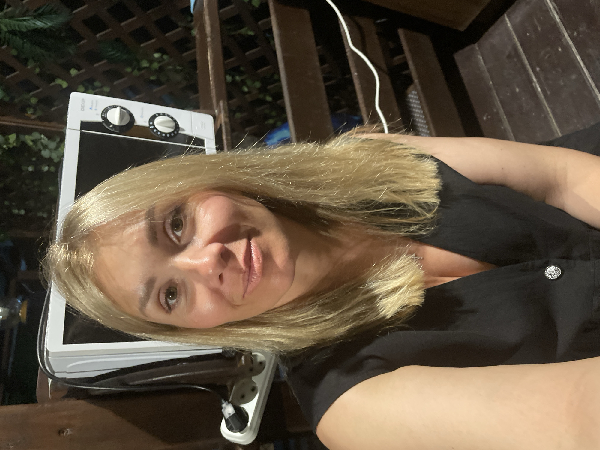 A woman with blonde hair and brown eyes is pictured in an outdoor setting. She is wearing a black top. Behind her, a white microwave oven is visible, along with a power strip and some wooden fencing. The lighting suggests it is evening or night.
