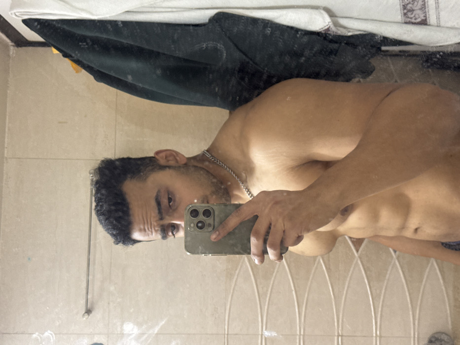 A man with dark, curly hair and a silver chain stands shirtless in a bathroom, taking a selfie in the mirror with an iPhone. The mirror shows a reflection of his muscular torso and abs. The bathroom has light-colored tile walls, and a towel hangs in the background, creating a cluttered yet intimate setting.
