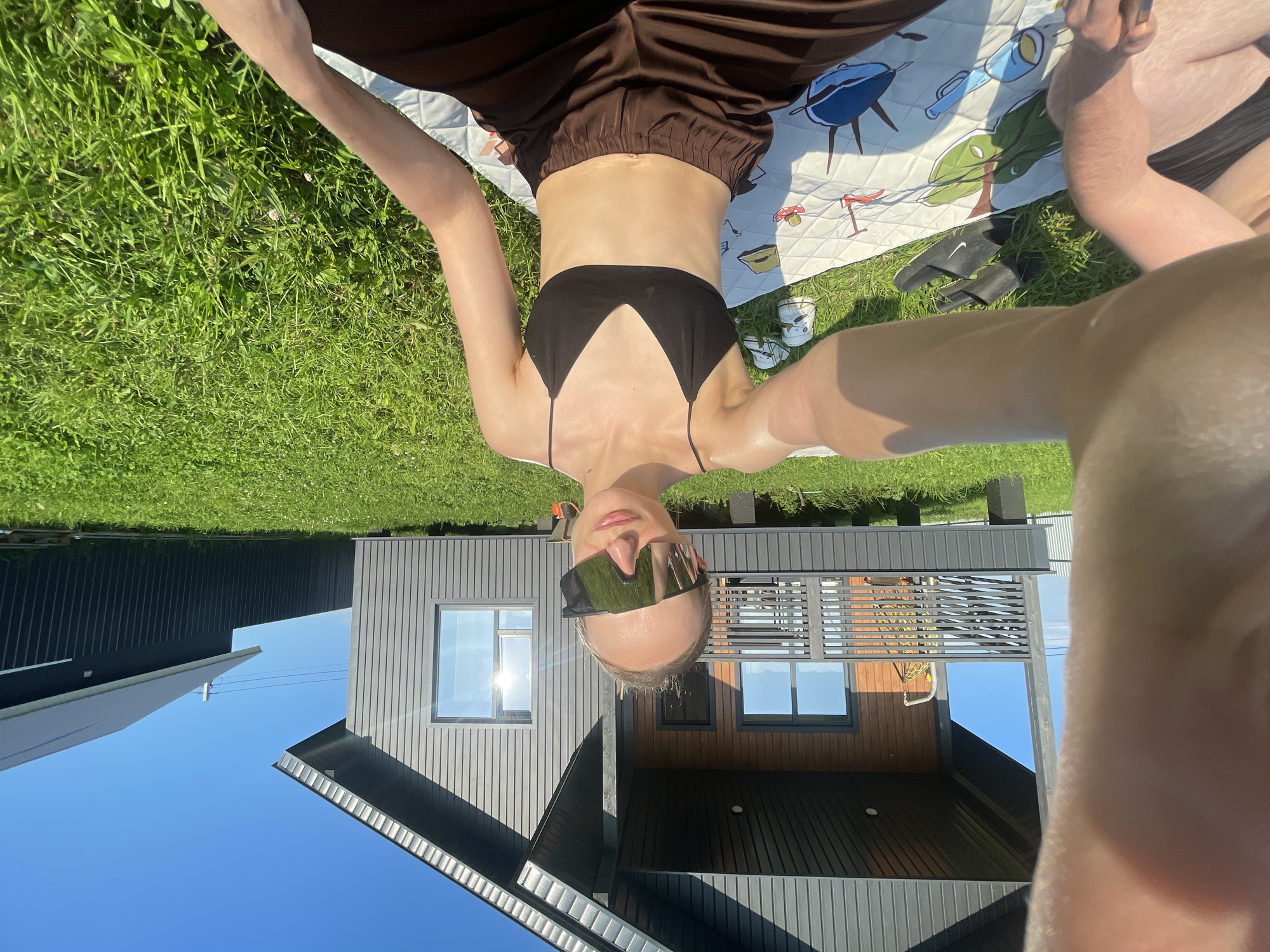 A person is lying on a picnic blanket on green grass, with their arms outstretched. They are wearing a black bikini top and brown pants. They have on mirrored sunglasses that reflect the sky and buildings. The background shows a modern house with dark siding and large windows. The sky is clear and blue, suggesting it is a sunny day. The scene appears to be a leisurely activity, possibly a picnic or sunbathing, taking place in a residential area in Podshivalovo, Russia. The time of day is likely midday, given the bright sunlight and shadows.