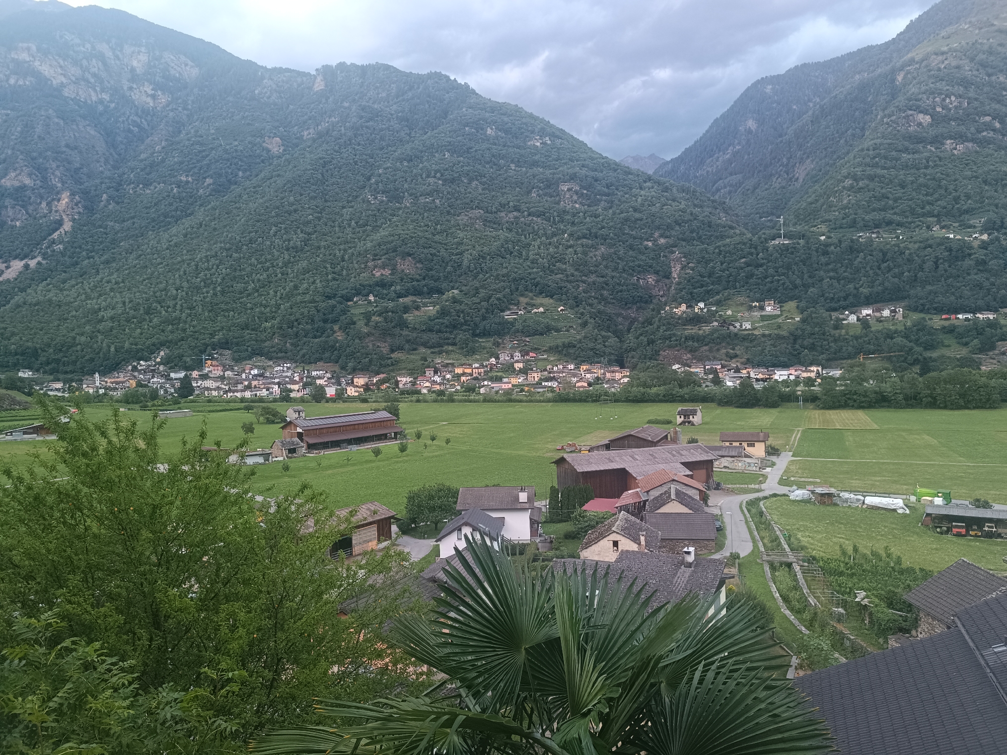 This is a high-angle, medium shot of a village nestled in a valley with mountains in the background, likely in Serravalle, Switzerland. The sky is overcast with dark clouds. The foreground is dominated by lush green trees and foliage. In the middle ground, a collection of houses and farm buildings with red and brown roofs are clustered together. A large, rectangular building with a brown roof and solar panels is visible in the distance. Beyond the buildings, expansive green fields stretch towards a town with more houses, and further still, the densely forested slopes of the mountains rise up. A road winds through the village. There are no people or animals visible in the image. The overall scene depicts a rural, mountainous landscape.