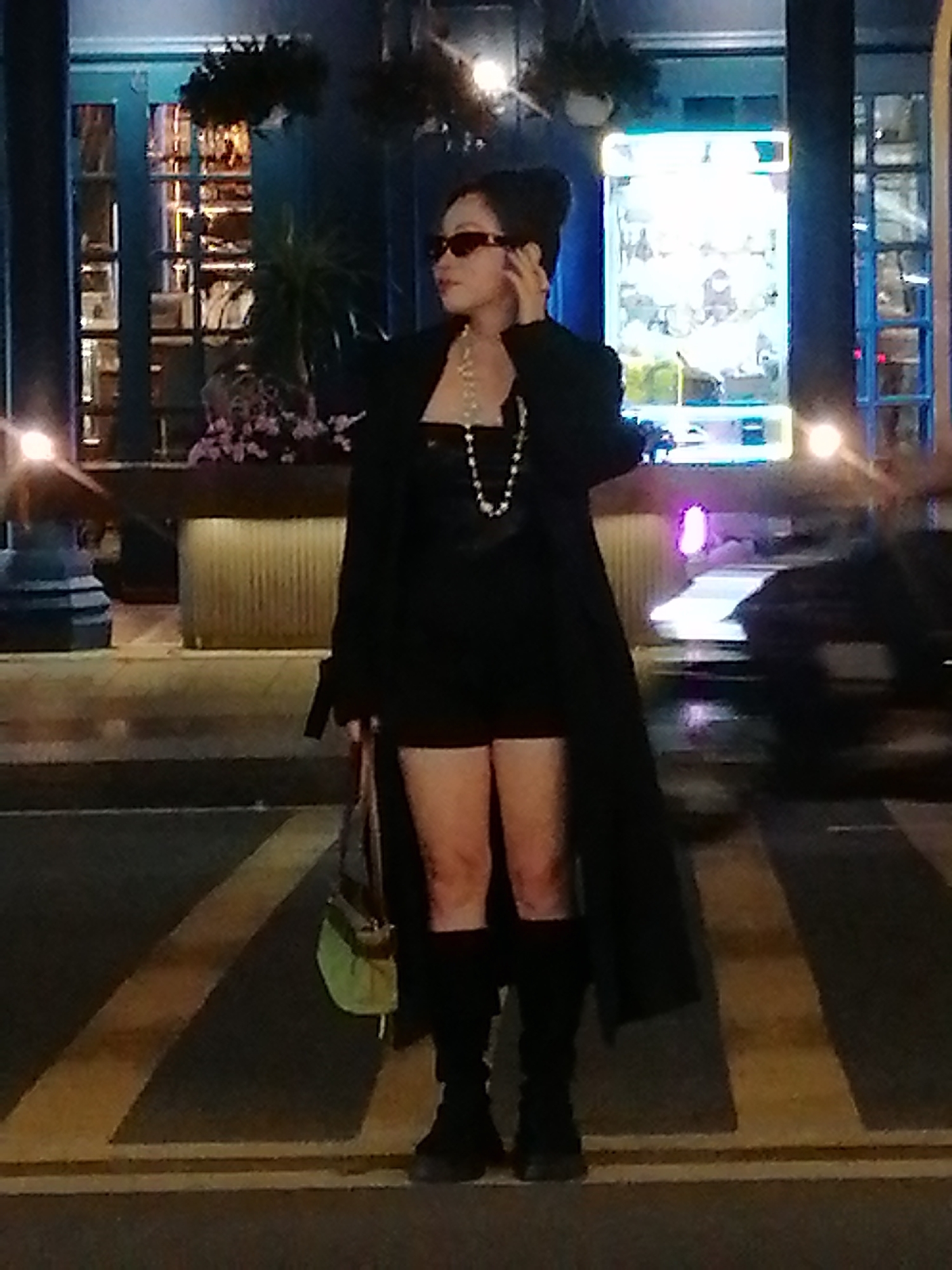 A woman stands on a crosswalk at night in Da Lat, Vietnam. She is wearing a black coat, a black strapless top, black shorts, and black knee-high boots. She has on sunglasses and a pearl necklace. She is holding a light green purse. In the background, there are buildings with glowing lights and some potted plants. A car is visible in the background, blurred by motion.