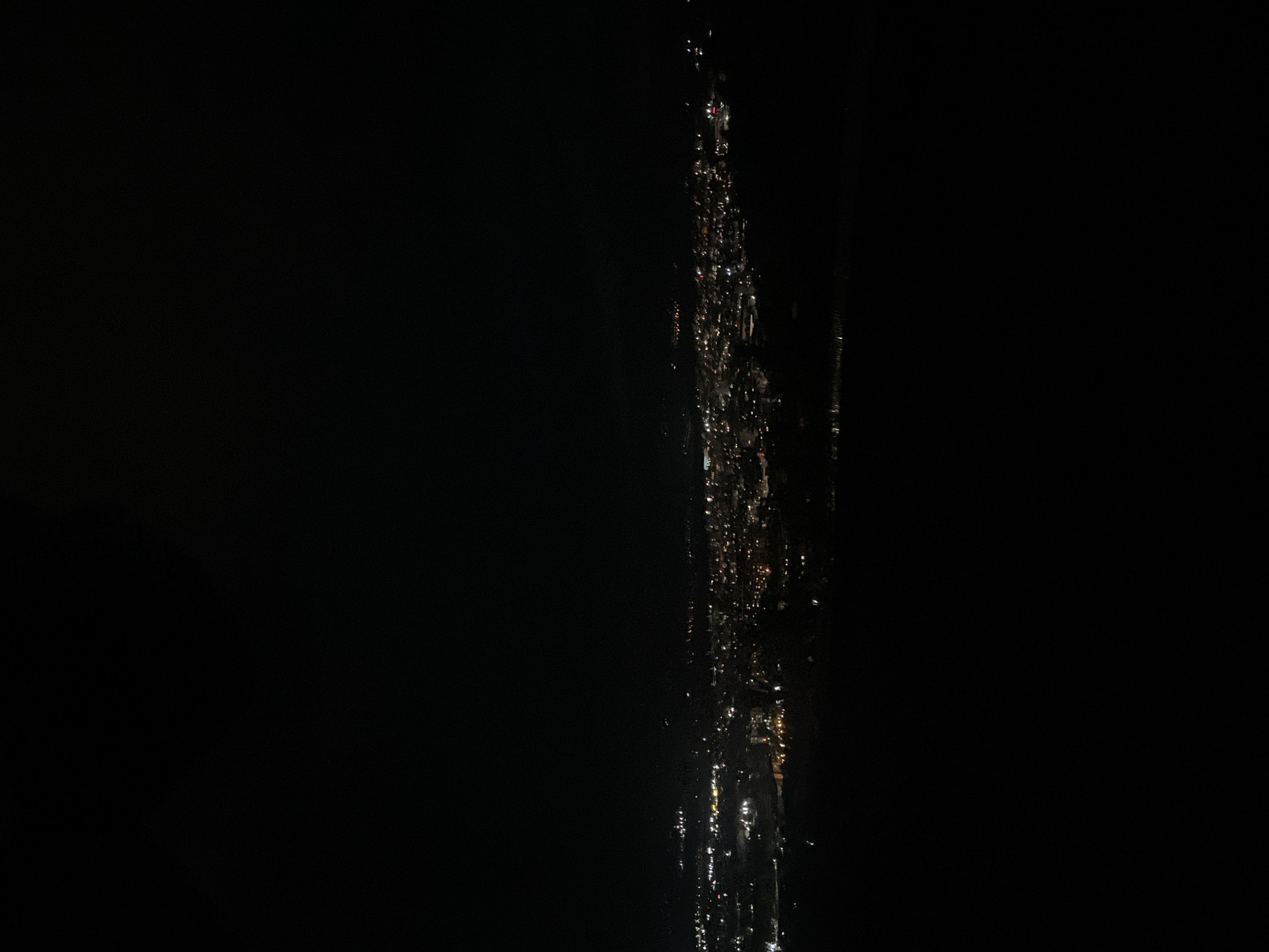 The image, taken at night, shows a dark environment with a narrow, vertical strip of blurred lights. The lights appear to be reflections on a wet surface. The image is too dark to ascertain any details about the location, people, or actions taking place.