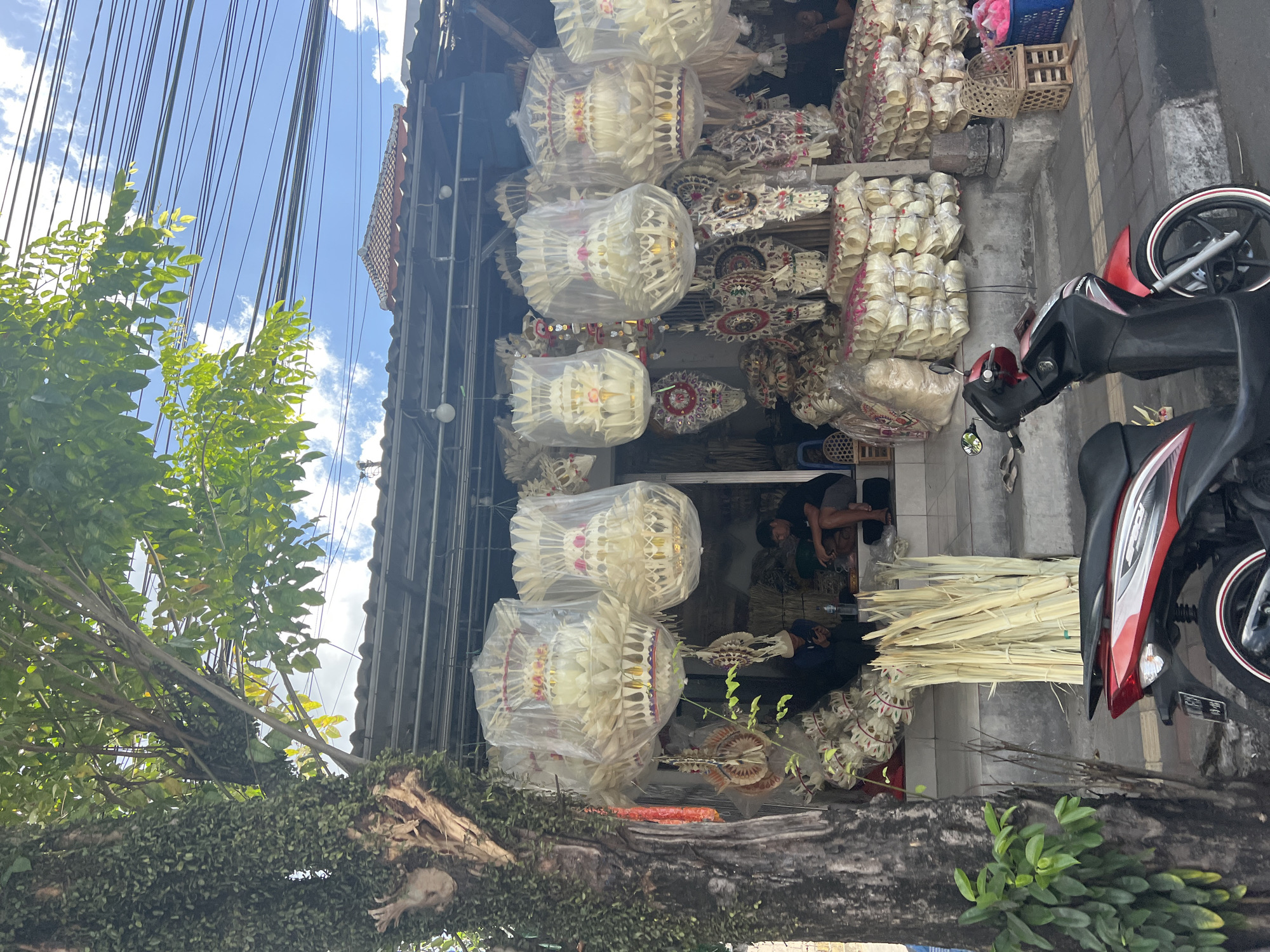 The image depicts a street-level view of a shop in Mengwi, Indonesia, selling traditional Balinese offerings. The shop front is filled with numerous intricately woven, layered offerings made of dried leaves, many of which are covered in clear plastic for preservation. Several larger, tiered offerings are suspended from the awning, also encased in plastic. Smaller decorative items, possibly also offerings or components thereof, are displayed hanging and stacked. In front of the shop, a large bundle of dried palm leaves is leaning against the wall. A red and black scooter is parked on the sidewalk in the foreground, partially obscuring a portion of the shop's offerings. Inside the shop, two men are visible, one seated and looking at a phone, and another sitting on a step. The shop has a simple awning and is situated next to a large tree with a trunk covered in dense green vegetation. The background shows a tiled roof and utility wires against a bright blue sky with scattered clouds. The ground is paved, with a curb and sidewalk.