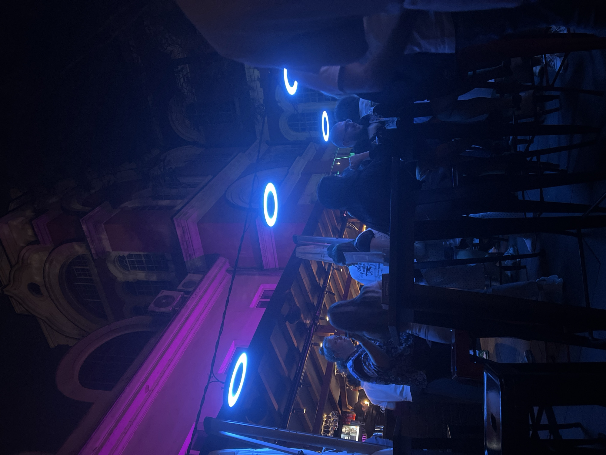 The image captures an outdoor bar or restaurant area at night in the city of Sofia, Bulgaria, situated in front of historical buildings. The scene is illuminated by a mix of neon and ambient lighting.
The left side of the upper background reveals a multi-story building with arched windows and classical architectural details, including a cornice line that is strongly lit with a magenta or purple hue. Below the cornice, two external air conditioning units are mounted on the building's facade. Further to the right, another older building with arched and rectangular windows, also displaying classical features, is visible, partially obscured by darkness and foliage.
The main subject area features a canopy structure from which several circular, blue LED lights are suspended, casting a cool glow on the patrons below. One additional, partially visible C-shaped blue light is seen further to the right.
Approximately 10-15 individuals are present within the frame, engaged in social activities typical of a bar setting. On the left, a man with long hair tied back, wearing a patterned shirt, is seen conversing with a woman. Behind them, a bar counter is visible with hanging glassware and a television screen displaying a woman's face. Further to the right, a man with glasses and a beard is smiling, looking towards an unseen individual. Other patrons are standing or seated at high tables with bar stools, many in silhouette or partial view due to the low light. Some people appear to be holding drinks. A menu or sign with "DUNHILL" printed on it is discernible on one of the tables in the midground. The foreground includes dark silhouettes of tables and chairs, and the right side is partially framed by the arm and shoulder of a person, likely the photographer. The overall ambiance is social and nocturnal, characterized by distinct artificial lighting and deep shadows.