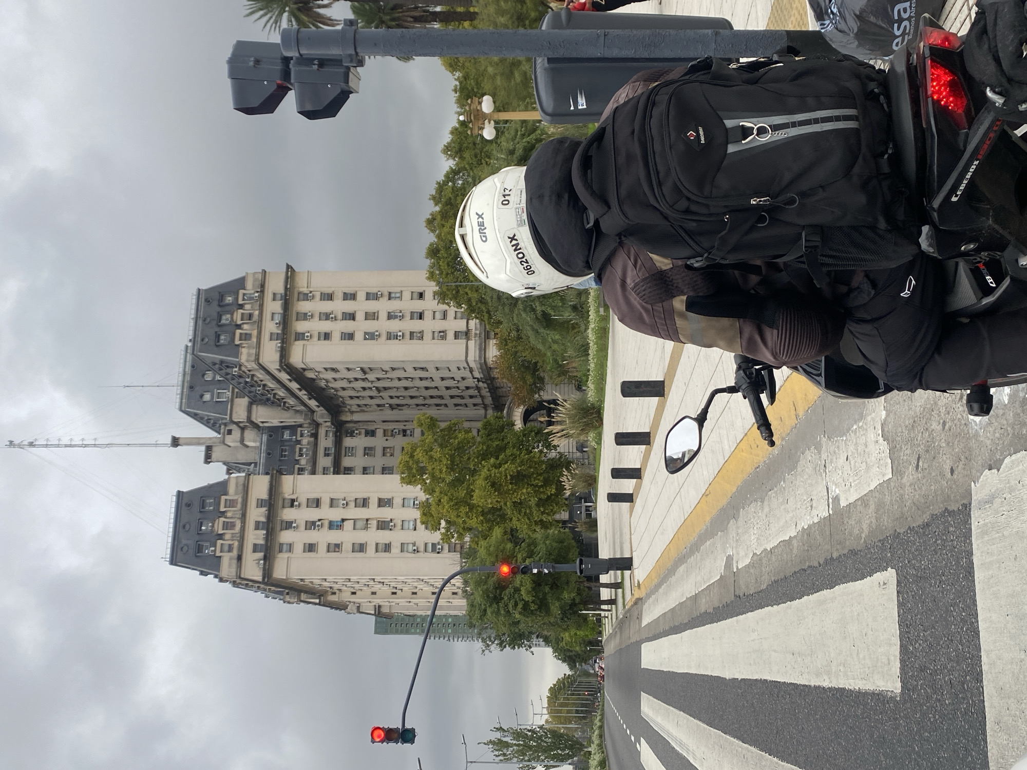 The photograph captures a motorcyclist stopped at a red traffic light on a city street in Buenos Aires, Argentina, under an overcast sky. The image is taken from behind the motorcyclist, who is wearing a dark jacket and a black backpack with reflective grey stripes and the brand name "BAGHERRY". A white helmet with text "GREX", "01?", and "XNOZ90" covers the rider's head.
The motorcycle's red tail light, a portion of its black body, and a side mirror are visible. The street ahead features a zebra crossing with distinct white and black stripes. In the background, imposing, multi-story buildings with classical architectural elements, numerous windows, and external air conditioning units line the street. Some buildings have dark, steep roofs, and one prominent structure in the distance displays a tall antenna or spire. Green trees are interspersed along the street and between the buildings.
Visible street furniture includes traffic light poles, a general street pole, and a black public trash bin. On a black bag situated near the motorcycle's rear, the words "Buenos Aires" and "esa" are clearly legible, confirming the geographical location. The motorcycle's tail section also shows partially readable text, appearing to be "CEBEROS 150R". The scene depicts a typical urban moment, paused by traffic.