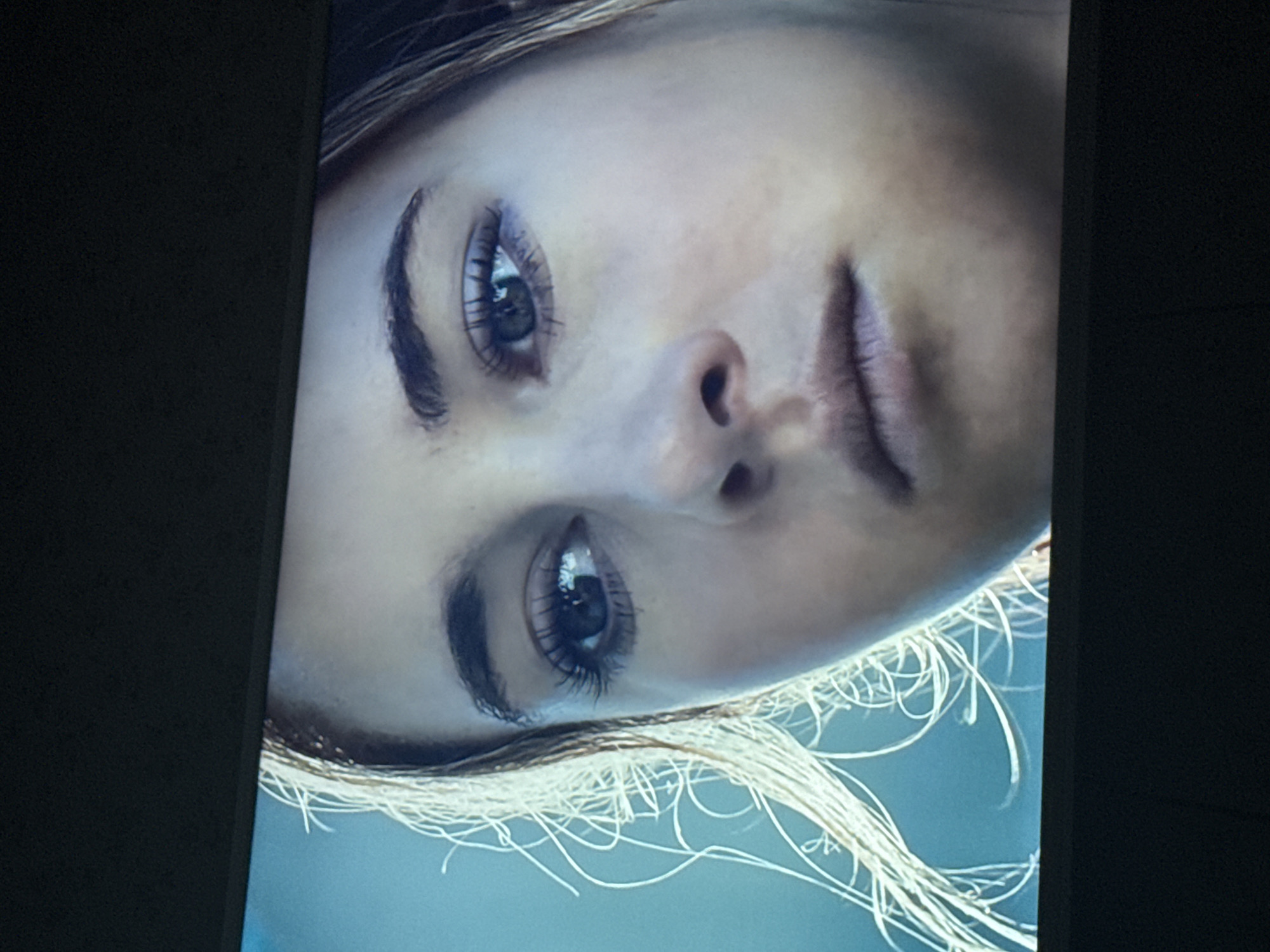 The image is a close-up shot of a woman's face, likely a still from a screen or poster, as indicated by the surrounding frame. She has fair skin, dark eyebrows, and light-colored eyes. The bottom portion of the image includes wisps of blond or light-colored hair against a light blue background. It is not possible to ascertain if this is in Mytishchi, Russia or any other specific location, based solely on the visual information provided.