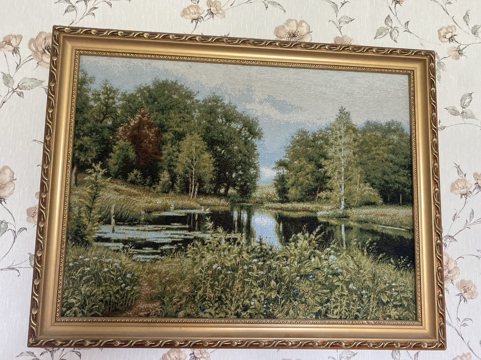 The image shows a framed tapestry depicting a landscape with a body of water surrounded by trees and vegetation. The tapestry is hanging on a wall with floral wallpaper. The scene features a serene lake reflecting the sky and trees, with reeds and wildflowers in the foreground. There are no people visible in the tapestry. The frame of the tapestry is ornate and gold-colored. The overall impression is one of a peaceful natural setting. The provided location in Vvedenka, Ukraine, is not verifiable from the image itself.