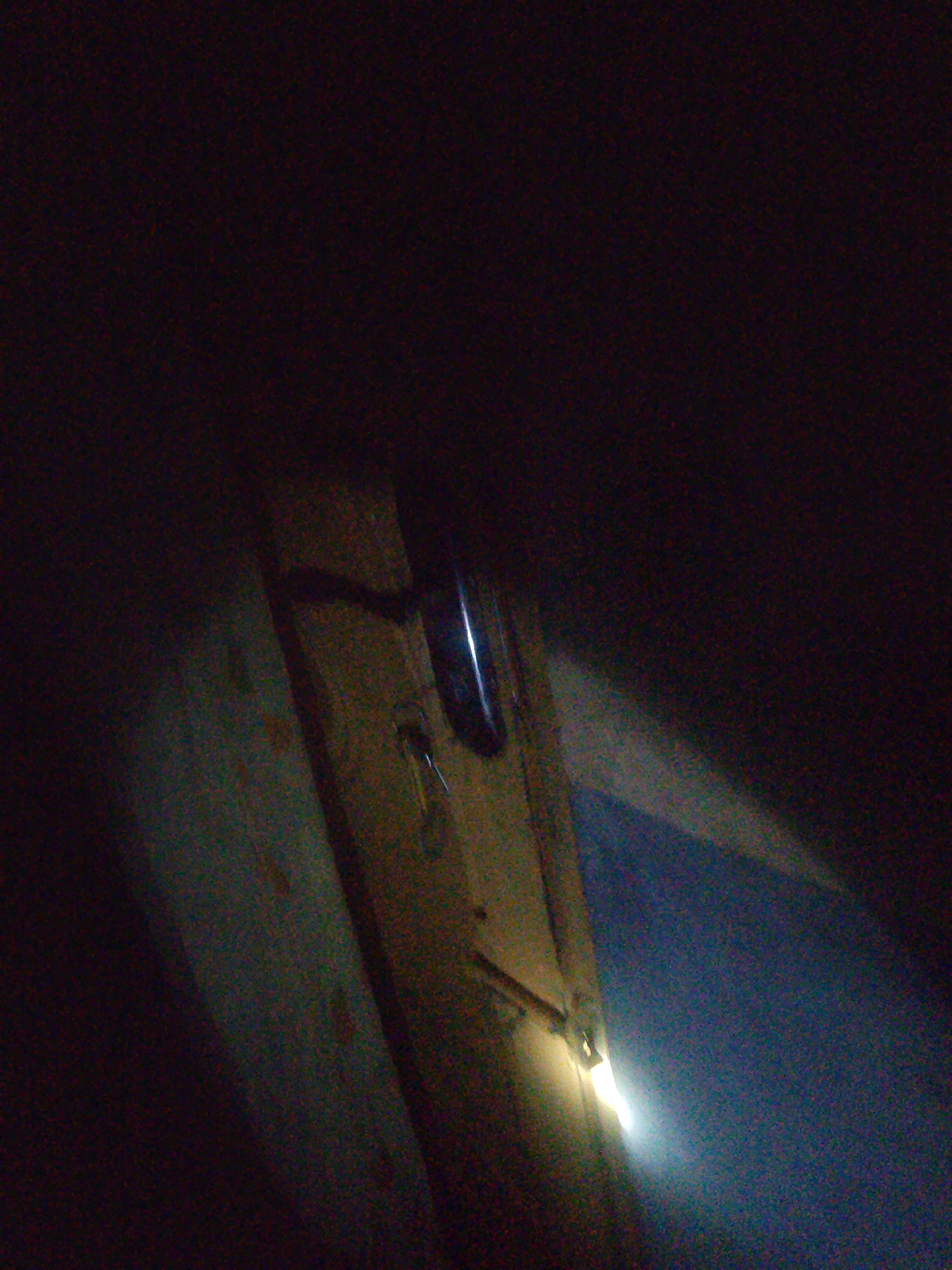 The image depicts a dimly lit, indoor scene. The primary subject is a wooden surface, possibly a shelf or a wall, with a metal latch or handle attached. A bright light source, likely a flashlight or a lamp, is illuminating the area from the lower right, casting a strong glare and creating a halo effect. The background is largely dark and indistinct, suggesting a confined space with minimal ambient light. The overall impression is one of a rustic or unfinished environment. The scene appears to be a close-up shot, focusing on the details of the latch mechanism and the texture of the wood. There are no people or other discernible objects in the image. The presence of the light source suggests an attempt to provide illumination in a dark setting.