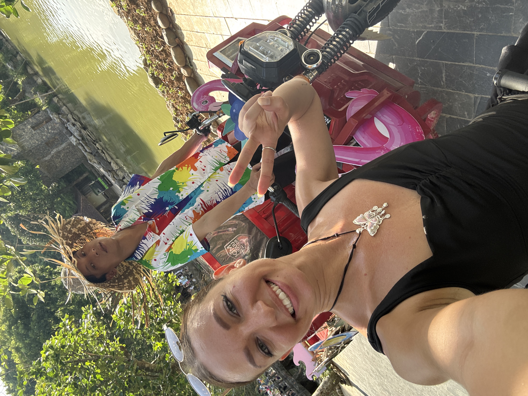 Two people are on a red electric scooter next to a body of water. The person in the foreground is a woman with a black top and a butterfly necklace, smiling and holding up two fingers. The person behind her is a man wearing a colorful shirt and a straw hat. The scooter is decorated with pink flamingos. The background shows trees, a stone building, and a body of water, suggestive of a park or resort. The scene is brightly lit, indicating it is daytime. This location is identified as Nam Hoa Lư Ward, Vietnam.