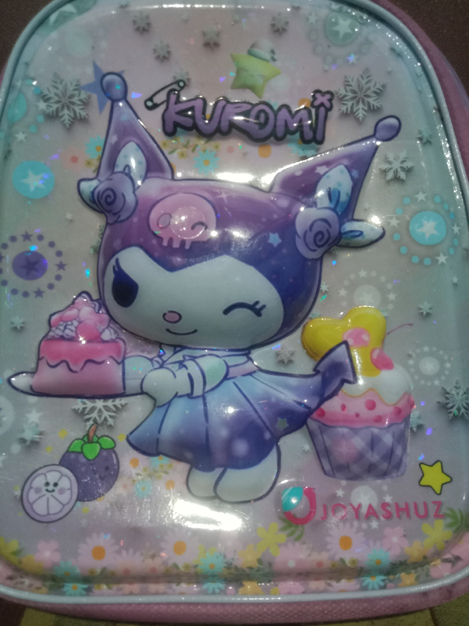 The image shows a close-up of a purple and white backpack decorated with the character Kuromi, holding two cupcakes. The backpack has a glossy finish and is adorned with stars, snowflakes, and flowers. A brand name, "JOYASHUZ," is visible at the bottom right of the backpack. The scene appears to be indoors, with the backpack placed on a surface that is not fully visible. There is no indication of the city of Kedungsari, Indonesia.