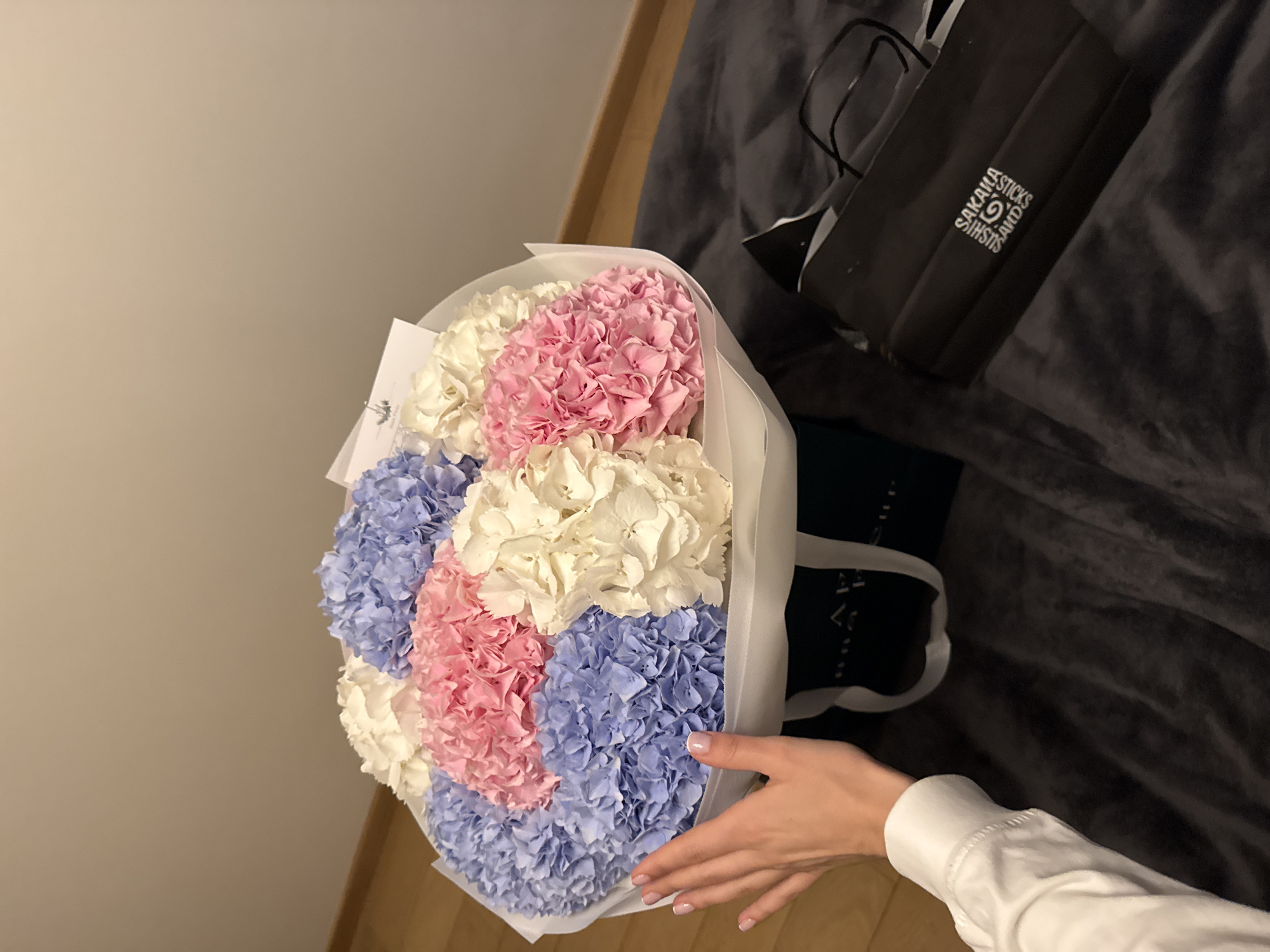 The image displays an indoor scene, likely in Warsaw, Poland, highlighting a person's hand interacting with a vibrant bouquet of hydrangeas. The large bouquet is artfully arranged in three distinct horizontal blocks of color: white, pale pink, and light blue hydrangeas, and is wrapped in translucent white paper. A small white card, partially visible within the bouquet, shows the text "FOR YOU."
A fair-skinned hand, clad in a white long-sleeved garment and featuring neatly manicured nails, gently touches the lower portion of the bouquet, suggesting the act of holding, receiving, or admiring the flowers. In the background, to the right, a dark grey textured fabric, possibly a blanket or duvet on a bed or couch, is visible. On this fabric rests a black paper gift bag with handles. The bag prominently displays white text that reads "SAKAWA STICKS AND SUSHI," along with a stylized logo incorporating these words, indicating a gift from a sushi establishment. The lighting in the scene is somewhat dim, characteristic of an indoor setting, possibly during the evening or with subdued artificial light. A light brown surface, likely a wall or wooden furniture, frames the left side of the composition. The overall impression is one of gifting or appreciation in a comfortable home environment.