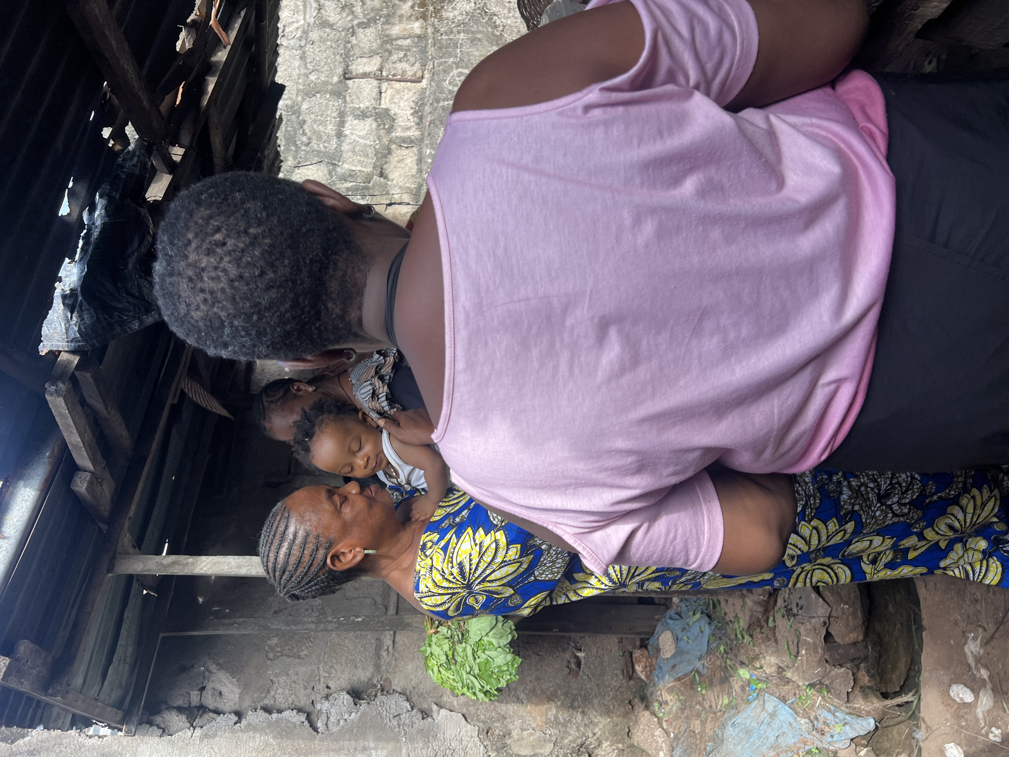 The image captures a moment in an outdoor setting. A woman in a patterned blue and yellow dress is holding a sleeping child. Behind her stands another individual in a pink shirt and black bottoms. The environment appears to be an impoverished area, with visible rough stone walls and deteriorated roofing. A bunch of green leaves is near the woman. The location is denoted as Unknown, Unknown.