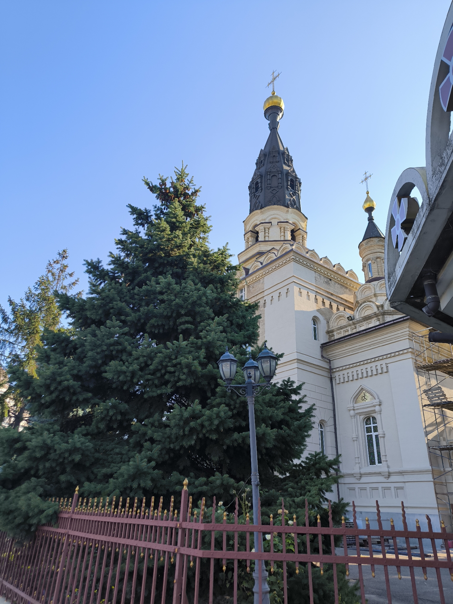 The image depicts a close-up view of a church building and surrounding elements, likely within the city of Mykolaiv, Ukraine. The architecture of the church is predominantly white with decorative beige accents, featuring multiple domes topped with golden crosses and a larger black bell tower with a golden dome. A tall, mature pine tree with dense green foliage dominates the left side of the frame, partially obscuring the church. In the foreground, a decorative reddish-brown metal fence with golden finials runs horizontally across the image, separating the viewer from the church and trees. A lamppost with three lights is positioned to the left of the tree. The sky is clear and blue, indicating fair weather. The scene suggests a daytime setting, possibly in the late afternoon given the shadows. There are no people visible in the image. Scaffolding is visible on the right side of the church building, suggesting ongoing construction or renovation work.